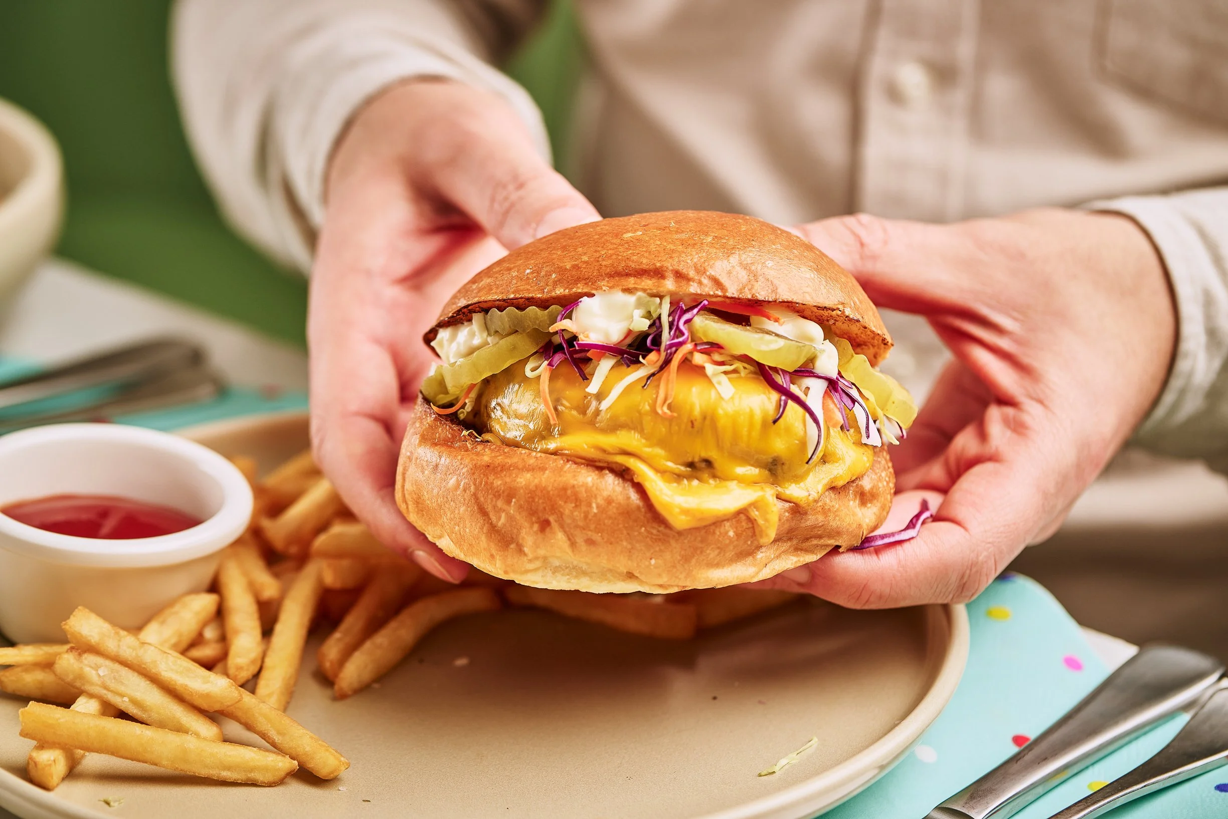 Hamburger with lettuce, tomato, cheese, and burger patty on a bun, served with French fries and ketchup, surrounded by colorful birthday party decorations, crayons, and a fruit bowl with cherry and yellow cherry tomatoes.