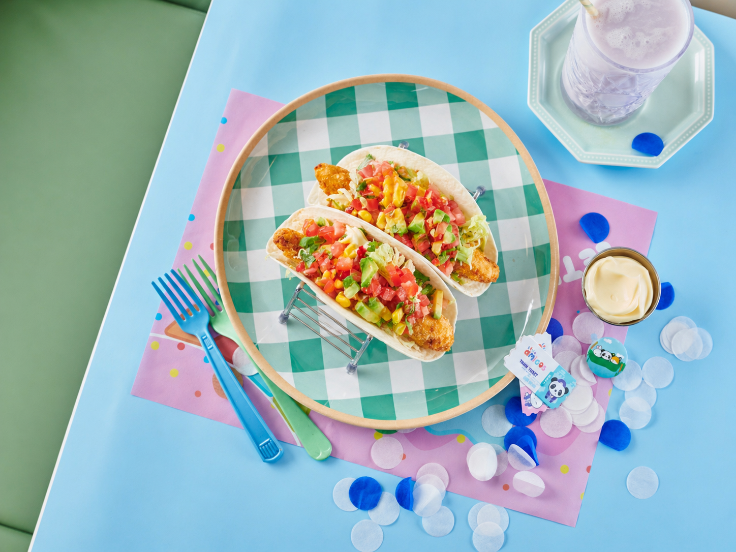 Three fish tacos on a white plate with shredded cabbage, diced tomatoes, and chopped herbs, served with a side of kale salad topped with nuts, on a yellow table with a fork and a bowl of mixed berries nearby.