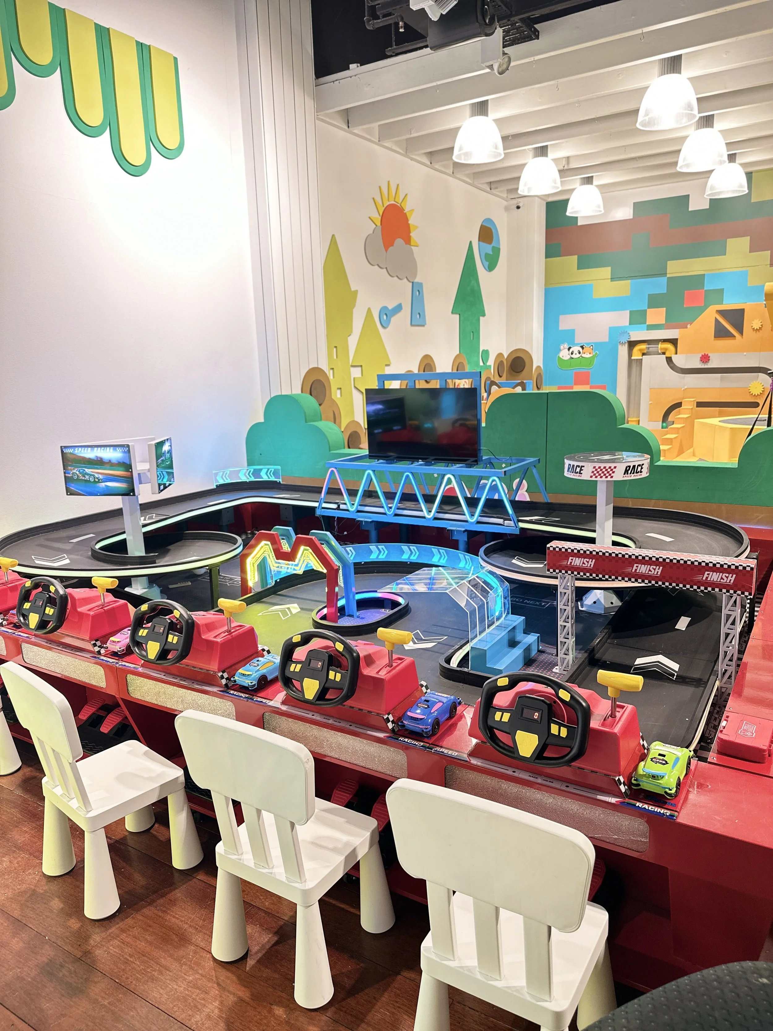 An indoor arcade with a small racing car game setup featuring four steering wheels and four small toy cars, chairs in front, colorful wall decorations, and a track with a finish line.