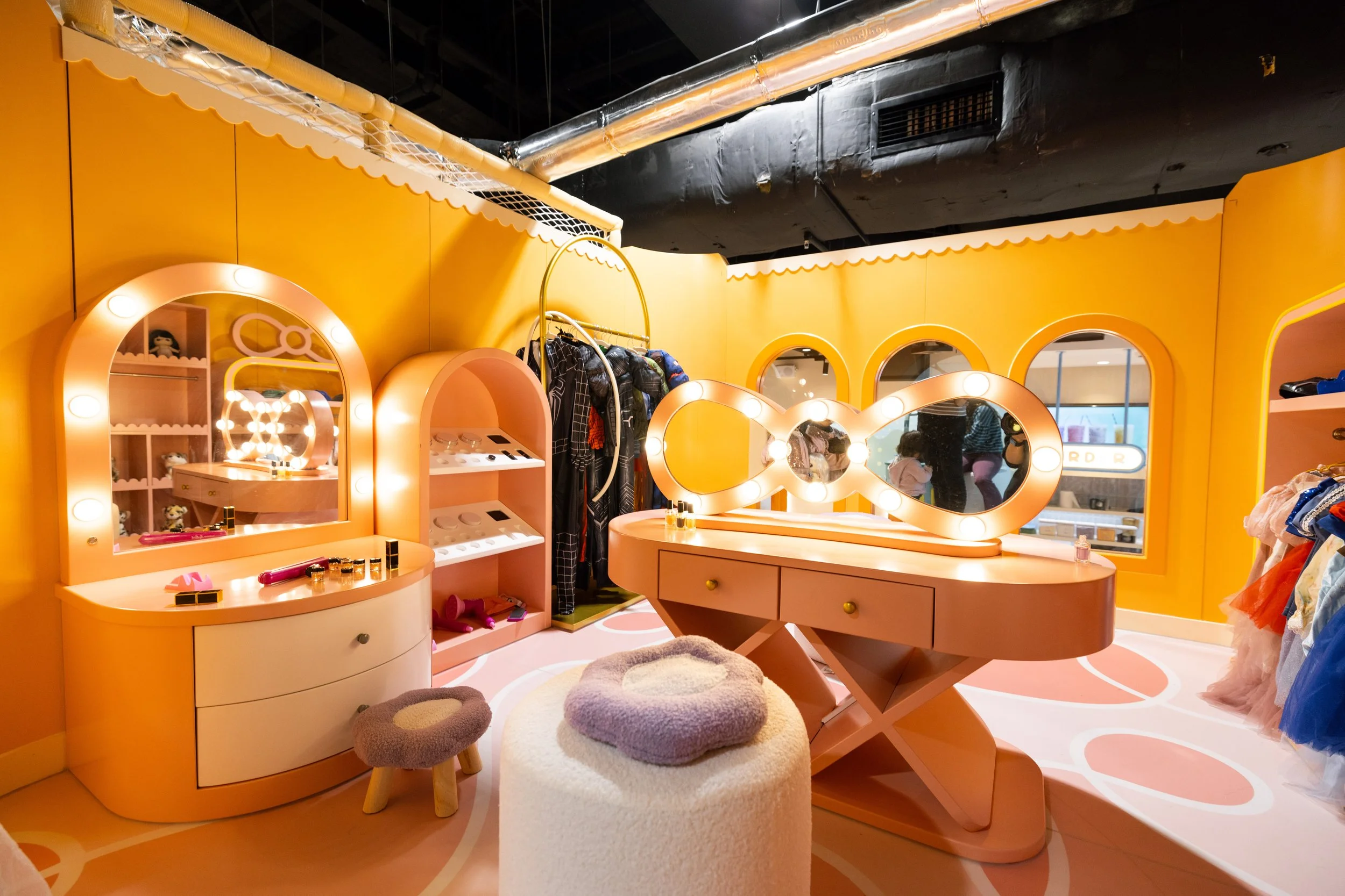 A children's dressing room with pink vanity tables, pink chairs, matching mirrors with round bulbs, and a clothing rack with costumes.