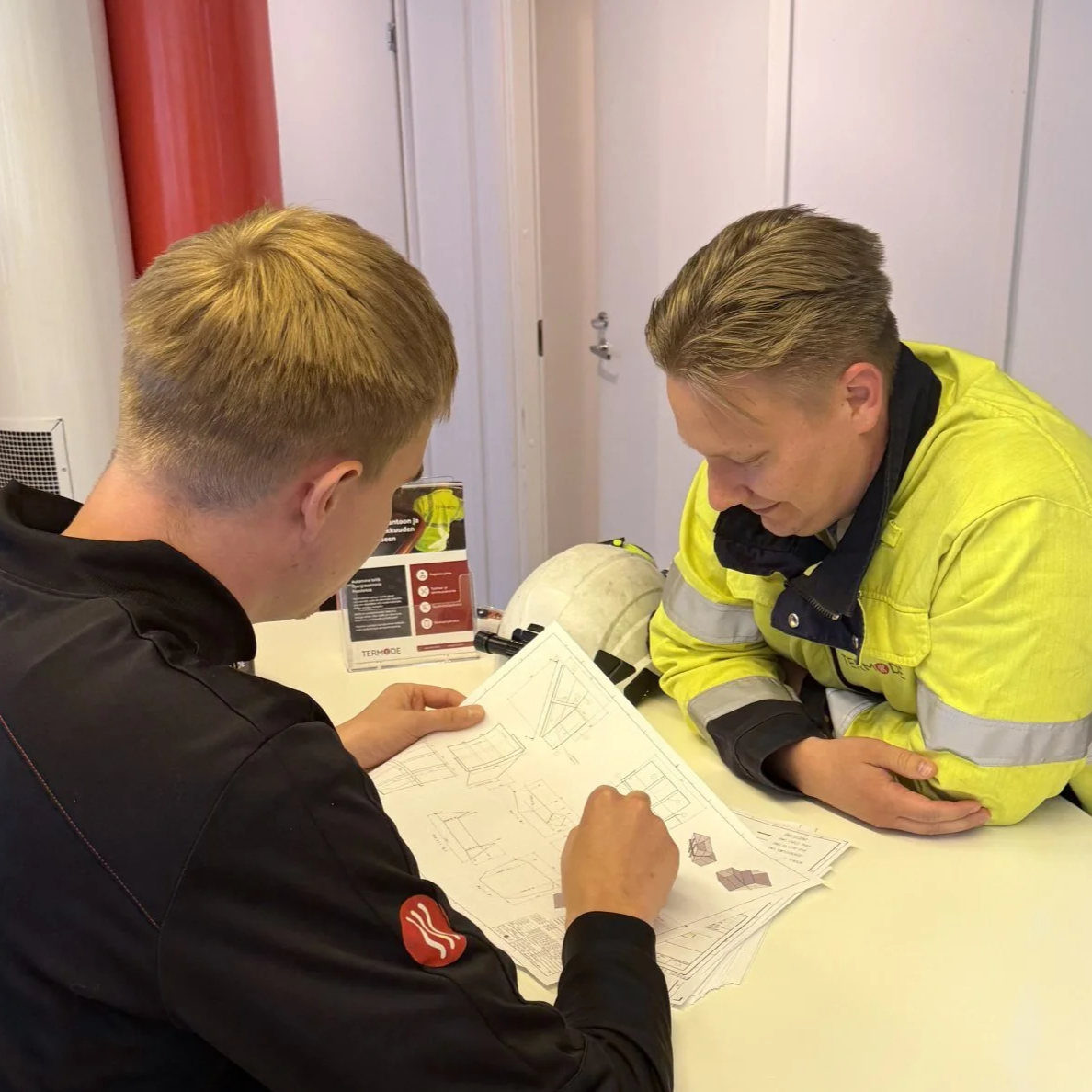 Kaksi työntekijää tarkastelee piirustuksia yhdessä, toinen suojavarusteissa.