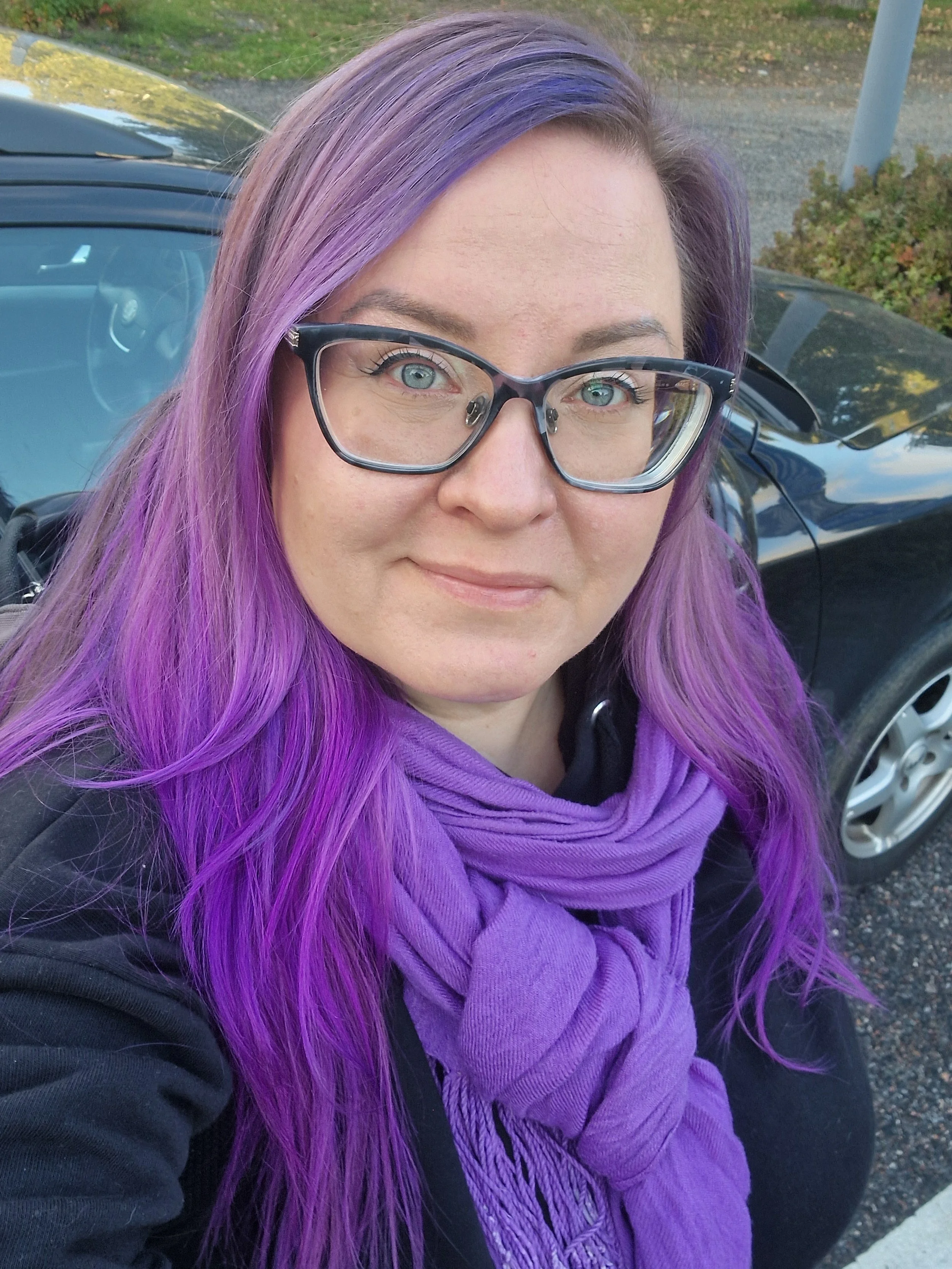 Nainen, jolla on violetit hiukset ja suuret kehykset, seisoo auton edessä ulkona, taustalla puita ja asfalttiteitä.