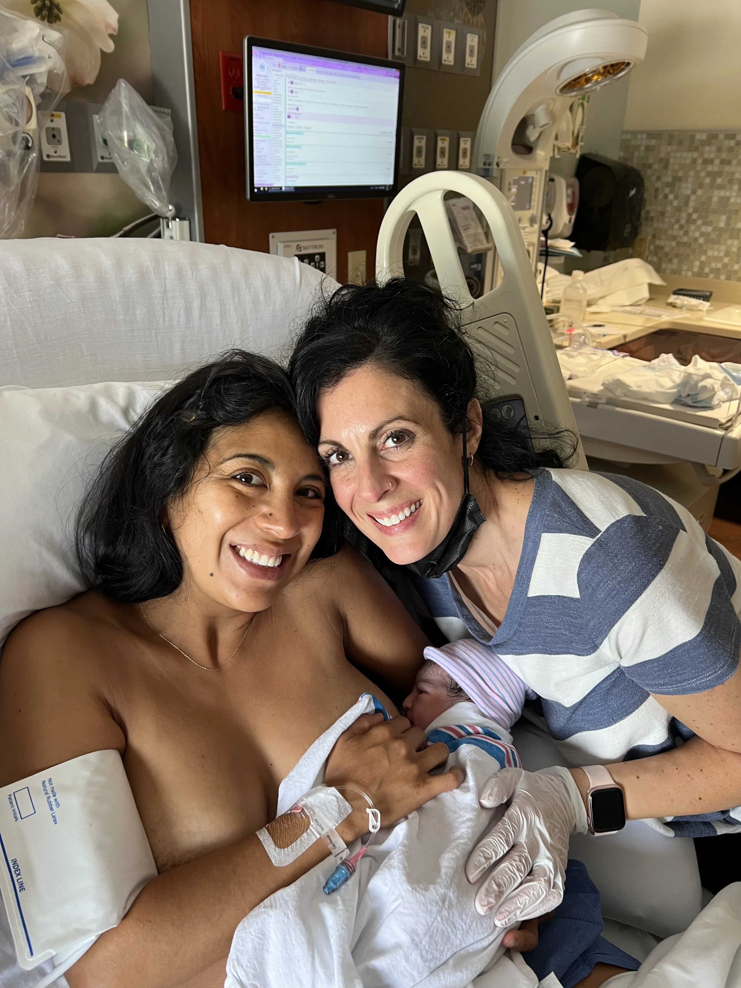 Woman in hospital bed holding newborn baby, smiling, with woman beside her wearing a striped shirt, all inside a hospital room.