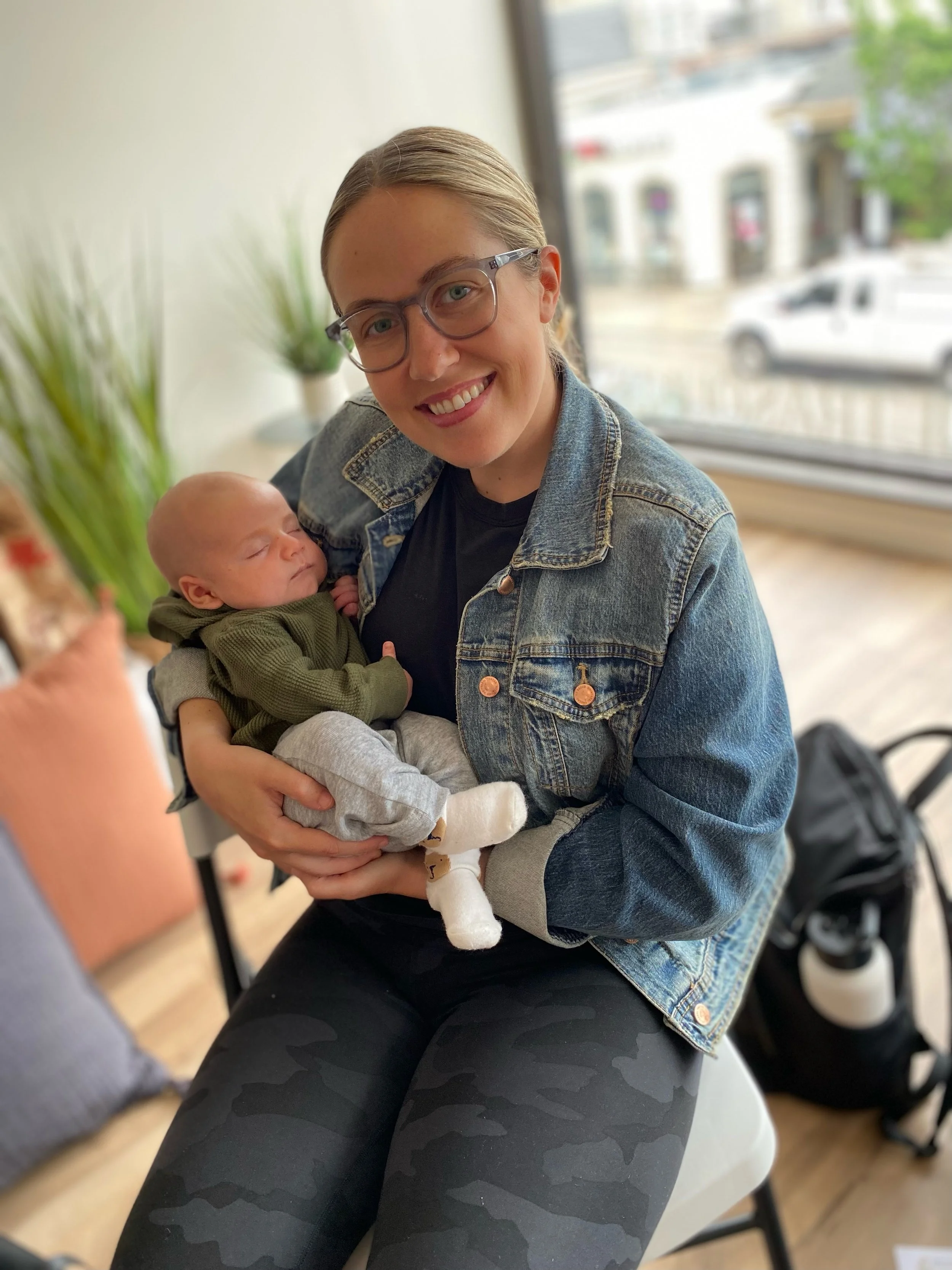 A woman with glasses and a denim jacket is holding a newborn baby in her arms. They are sitting indoors near a large window with a view of a street outside. The woman is smiling at the camera, and the baby is peacefully sleeping.