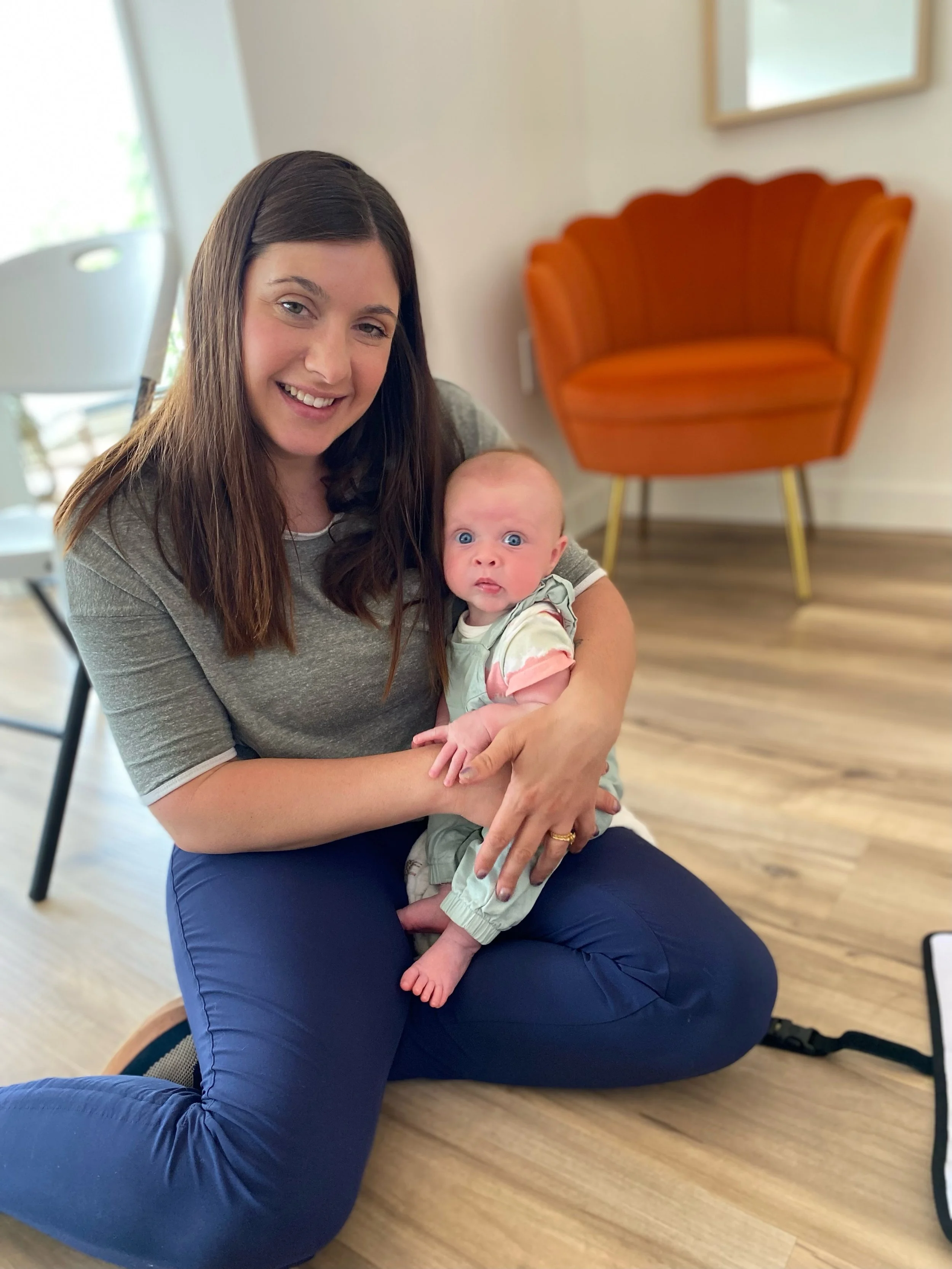 A woman with brown hair smiling while holding a baby with blue eyes, sitting on the floor of a room with wooden flooring, a white wall, an orange chair, and a window.
