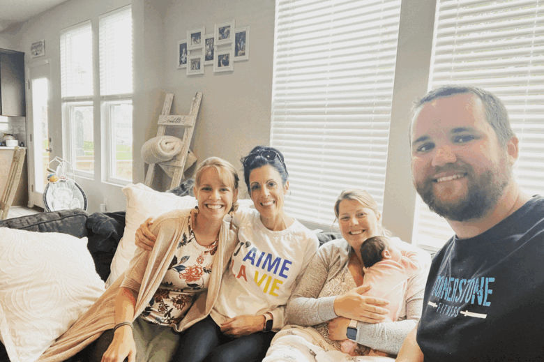 A group of five people seated on a couch in a well-lit living room, smiling at the camera; one woman is holding a baby, another woman and man are sitting close together, and the room features large windows, a ladder shelf, and framed photos on the wall.