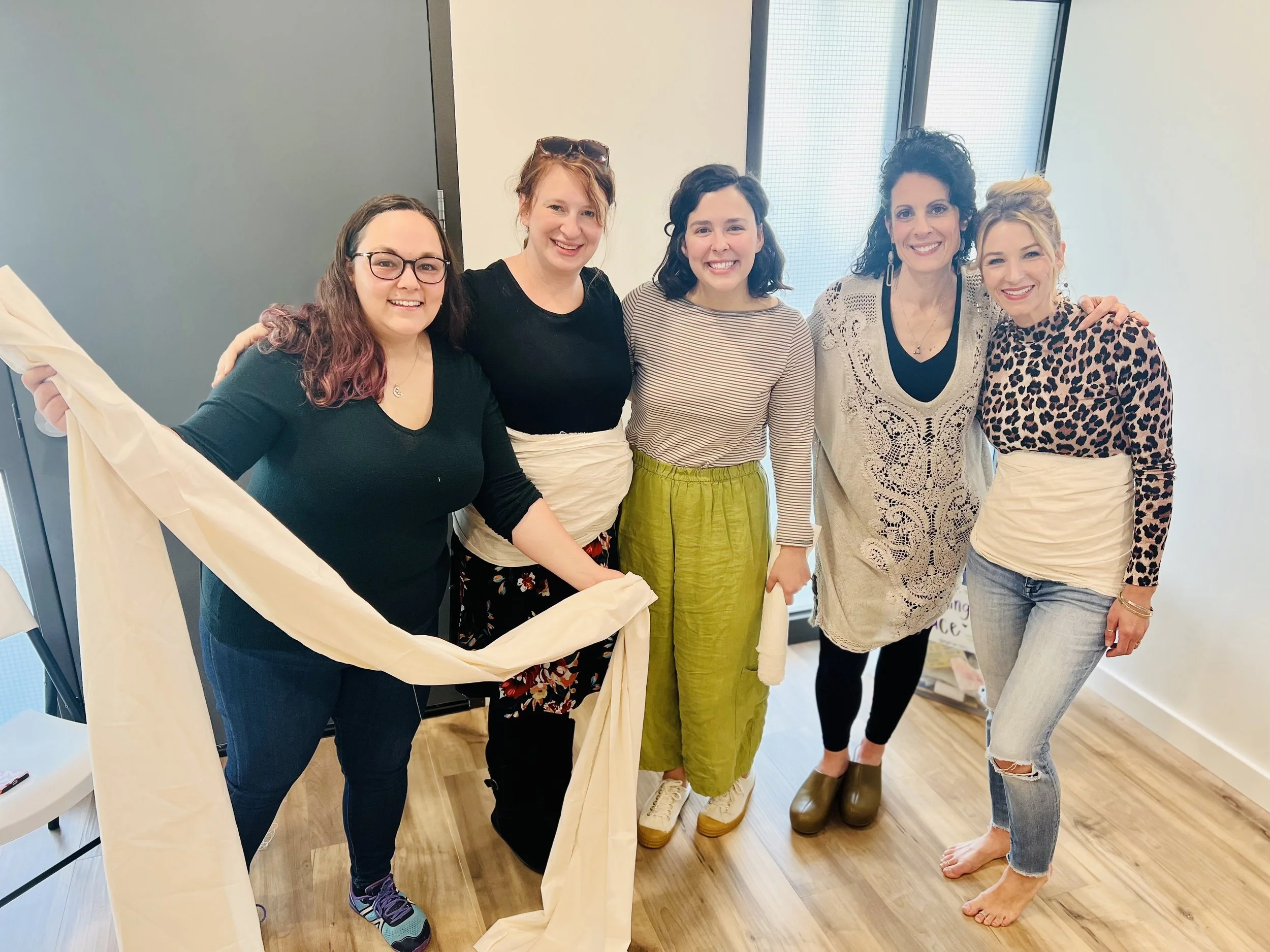 Five women standing in a row indoors, smiling for the camera. The woman on the far left is holding a long piece of fabric.