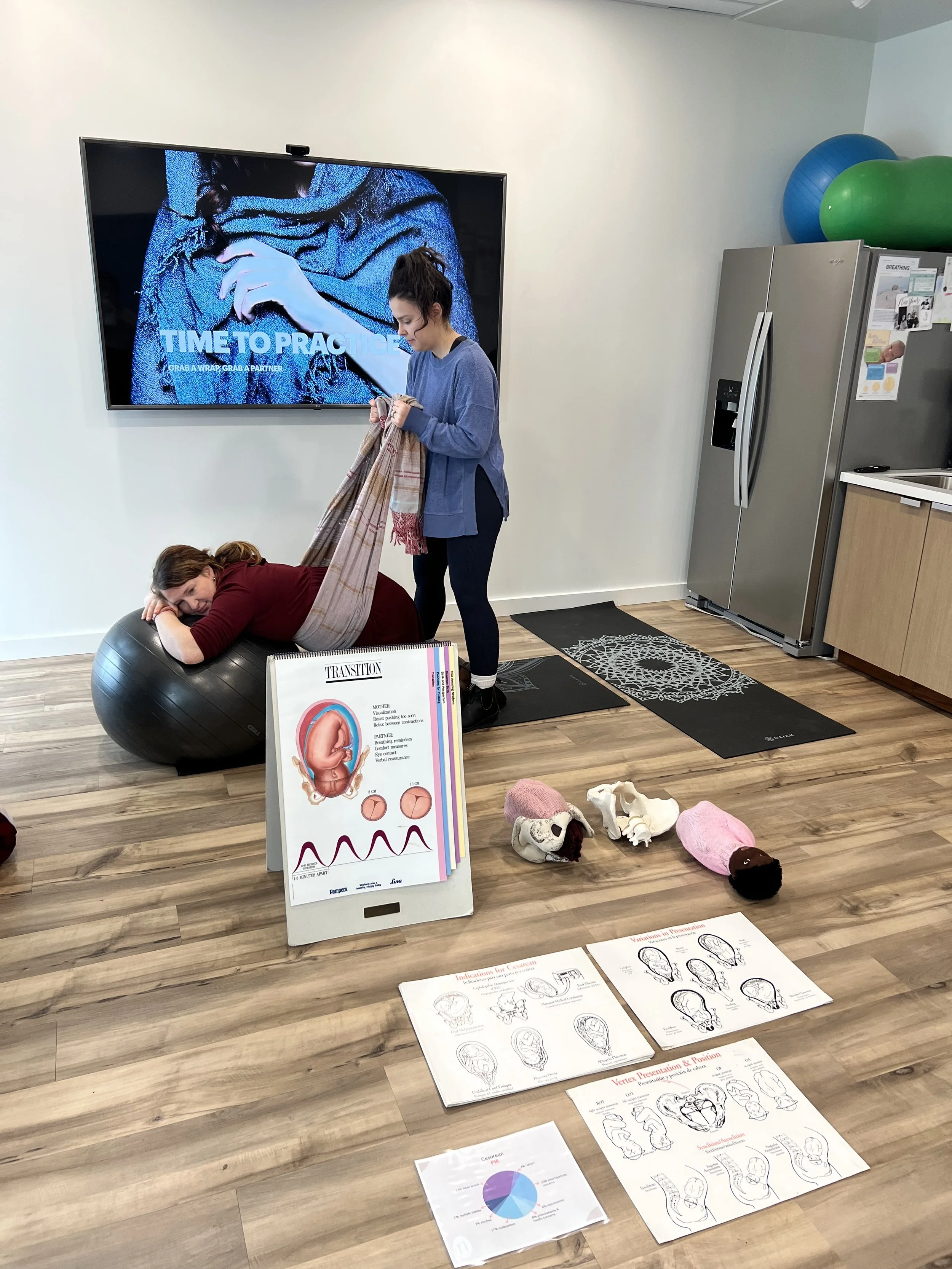 Two women in a room with educational materials on childbirth, including a poster, diagrams, and plush uterus models, with one woman lying on an exercise ball and the other holding a blanket.