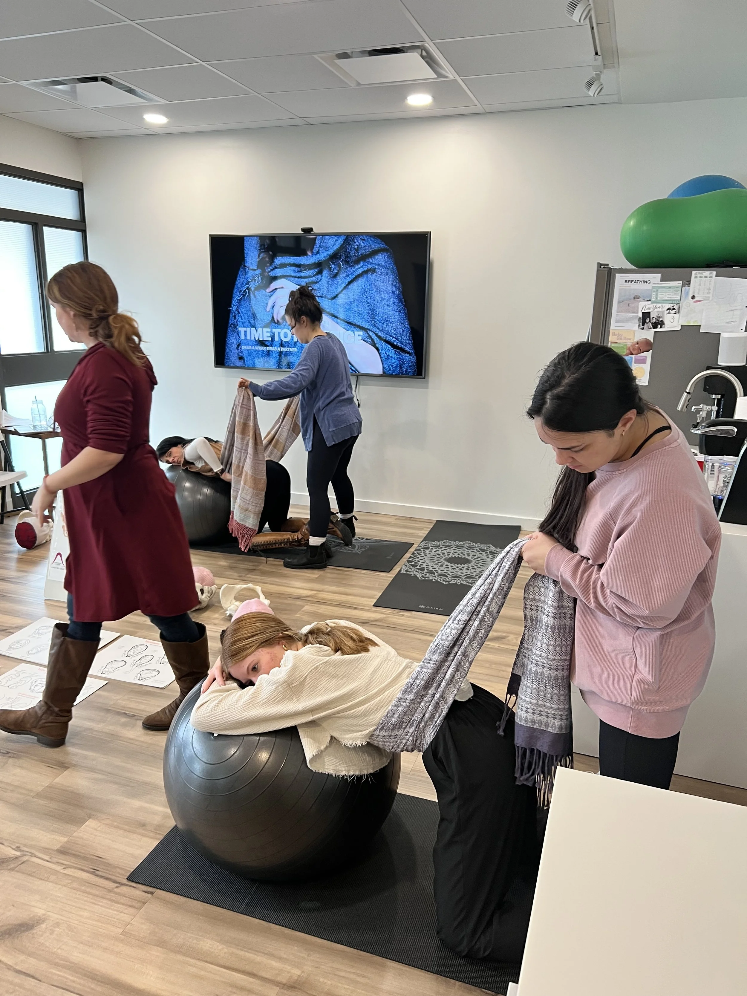 Women participating in a wellness class with exercise balls, some lying on their stomachs, others holding scarves, in a room with fitness mats, a screen, and workout equipment.