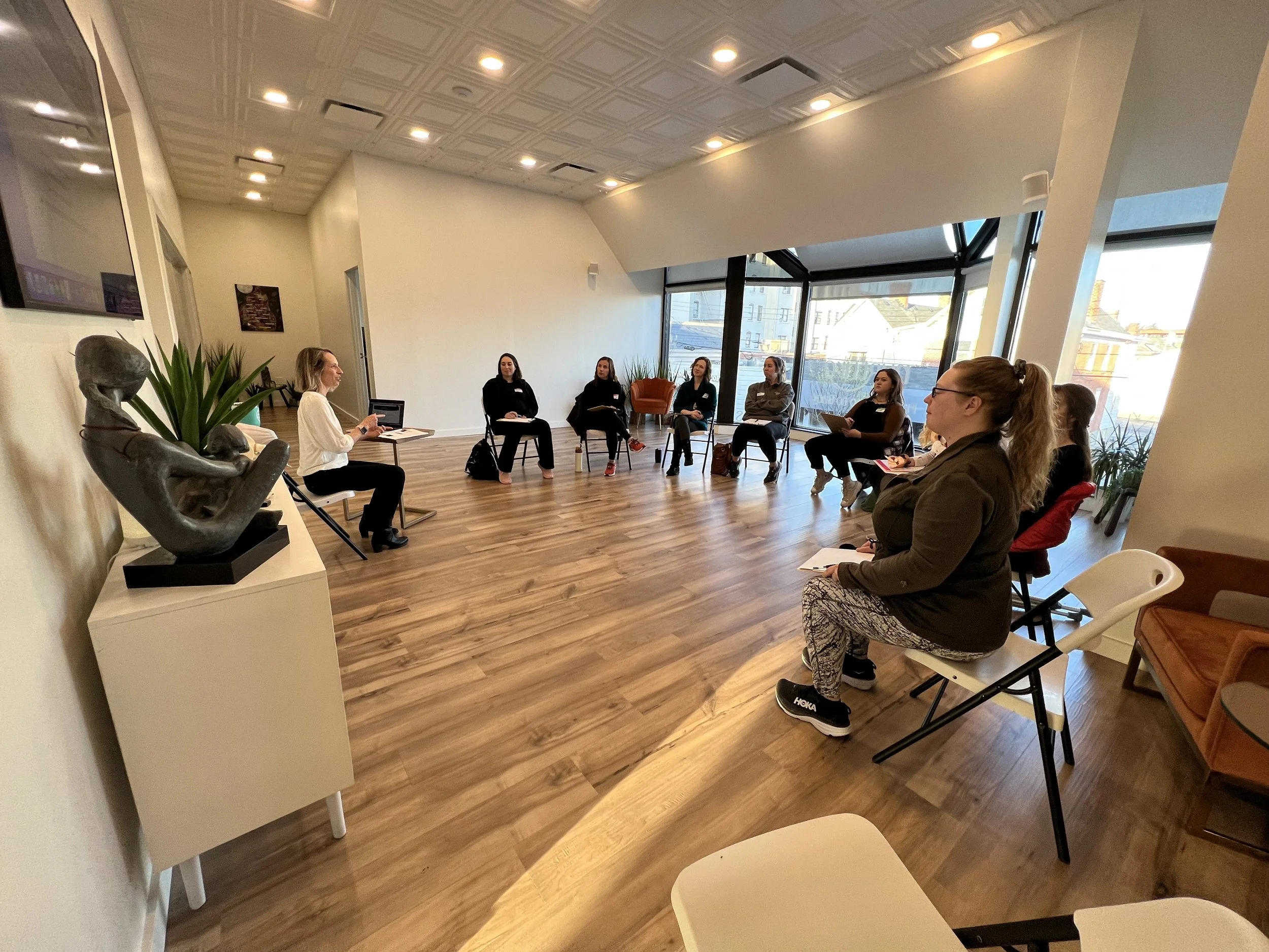 A woman leads a discussion in a seminar or workshop with seven women seated in a semi-circle in a bright, modern room with large windows, wooden floors, and sculptures on a white shelf.