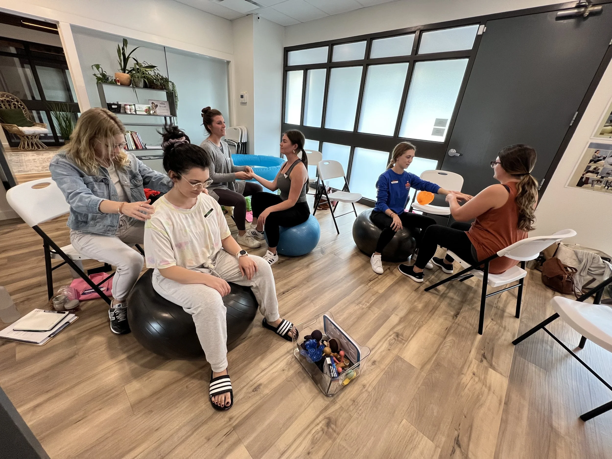 Women in a wellness class practicing seated stretching and relaxation exercises using exercise balls, with some holding hands and others receiving assistance, in a bright room with wooden floors and large windows.