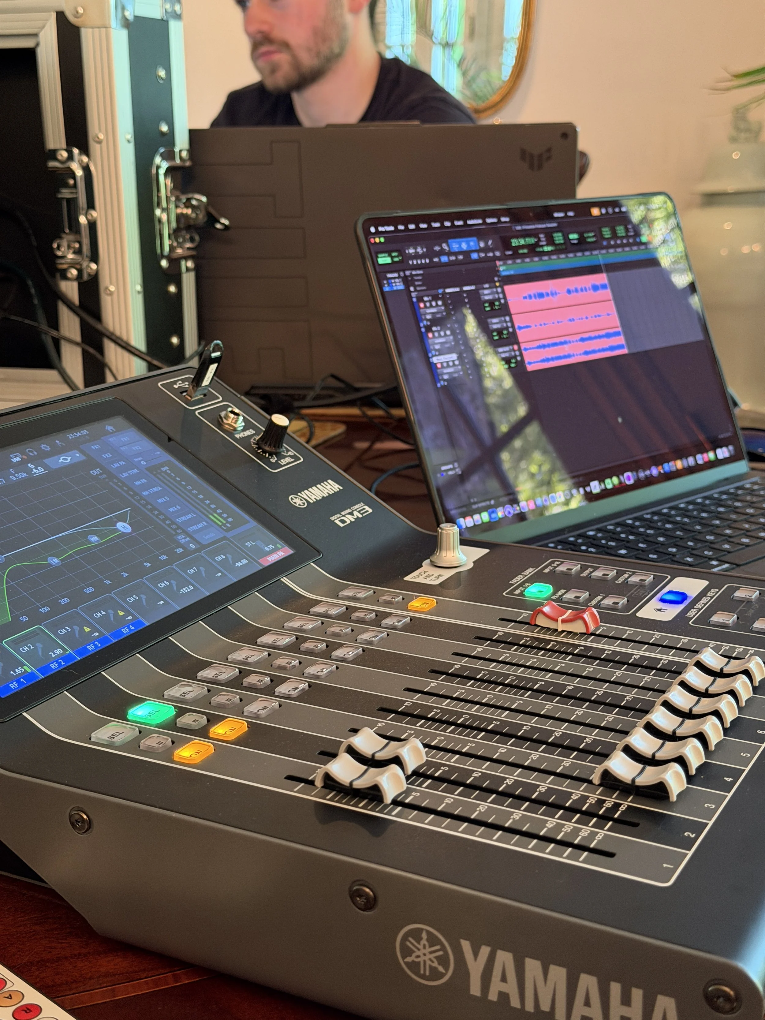 Music production setup featuring a Yamaha mixing console with dials, buttons, and sliders, a computer displaying audio editing software with waveforms, and a person in the background working on a computer.