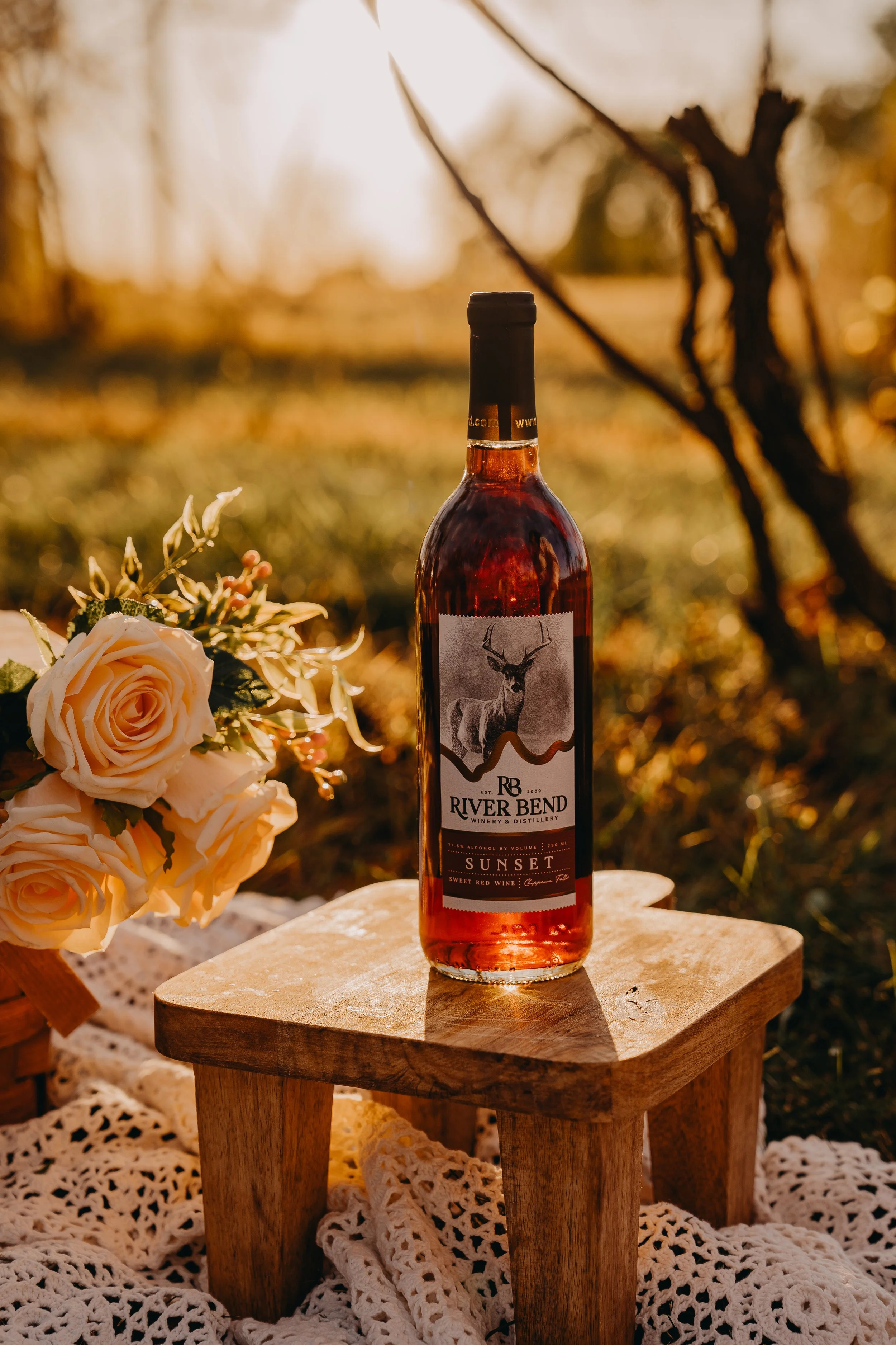 A bottle of River Bend Sunset red wine on a small wooden table outdoors during sunset, with white roses and a crocheted cloth nearby.