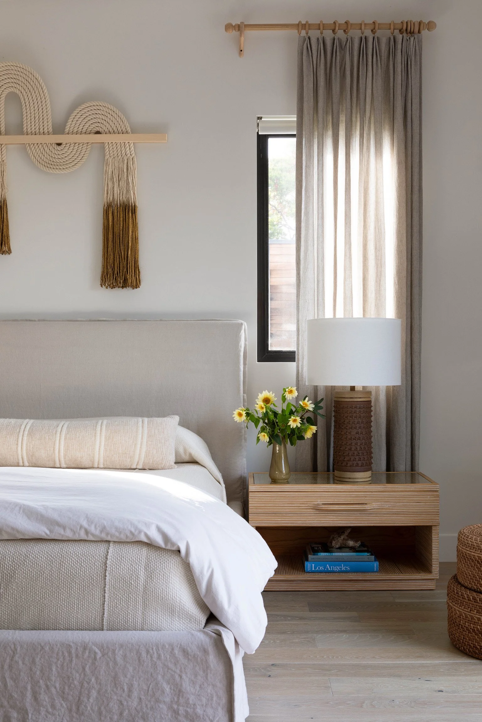 Los Angeles primary bedroom with custom drapery, woven wall art, warm wood nightstand and layered neutral textiles.