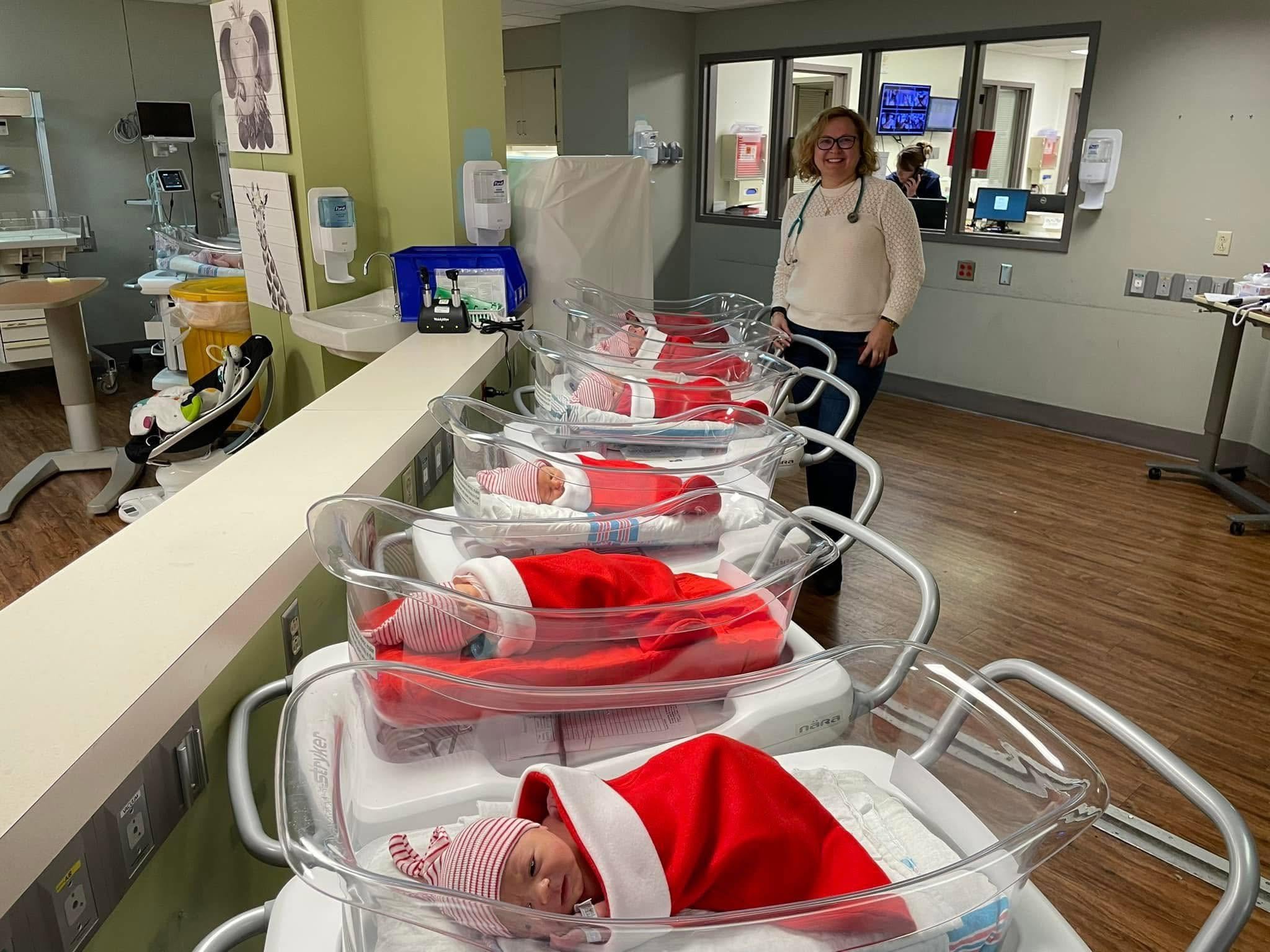 A hospital nursery with six newborn babies in clear plastic cribs, all dressed in red and white striped clothing and Santa hats. A smiling female healthcare professional stands beside the cribs, wearing glasses, a white sweater, and a stethoscope around her neck. Medical equipment and monitors are visible in the background.