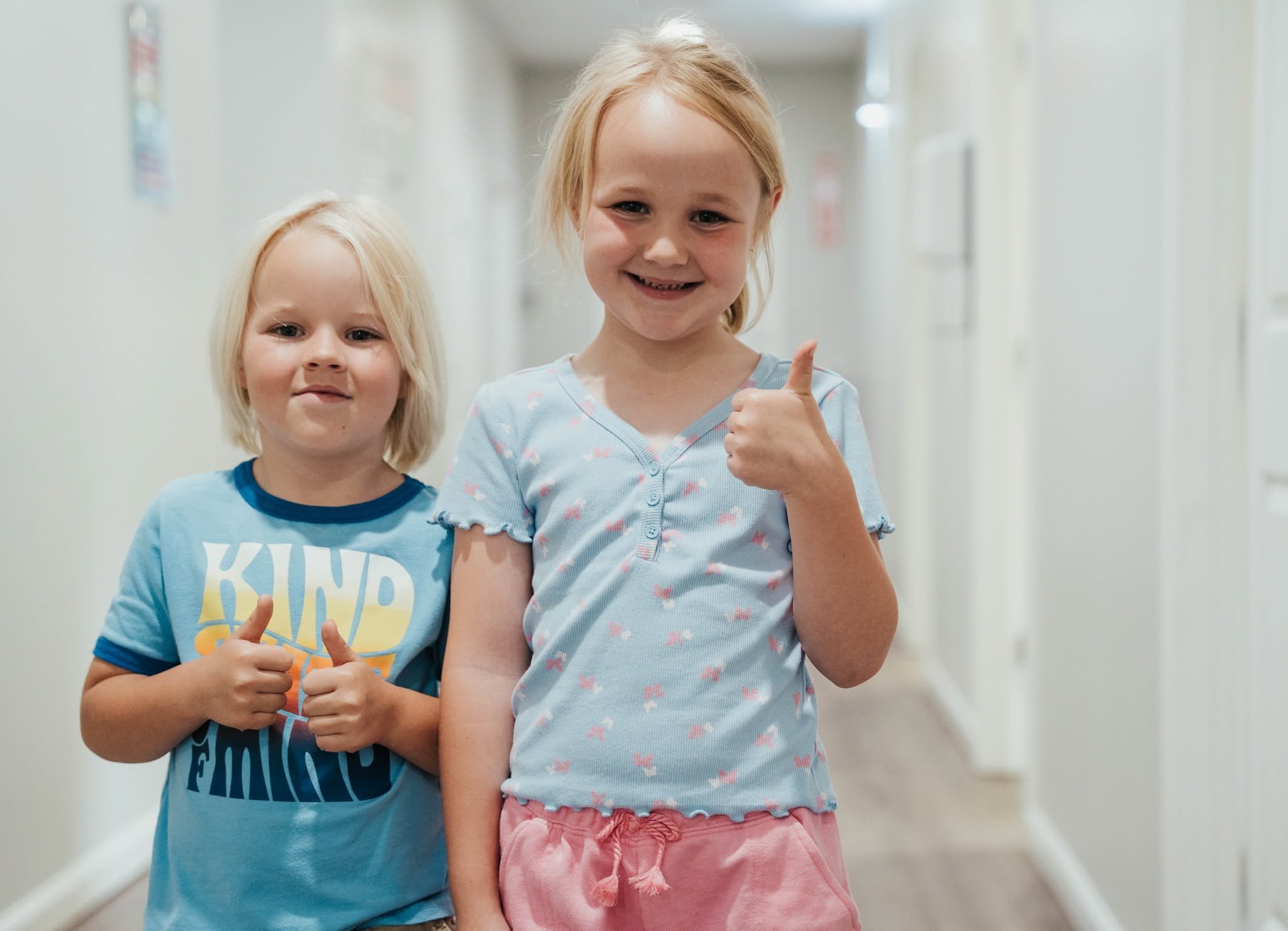 Two young girls standing in a hallway with white walls and doors, smiling at the camera. The girl on the right is giving a thumbs-up and wearing a light blue shirt with pink shorts. The girl on the left is making a peace sign with her fingers and wears a blue shirt with yellow and white lettering.