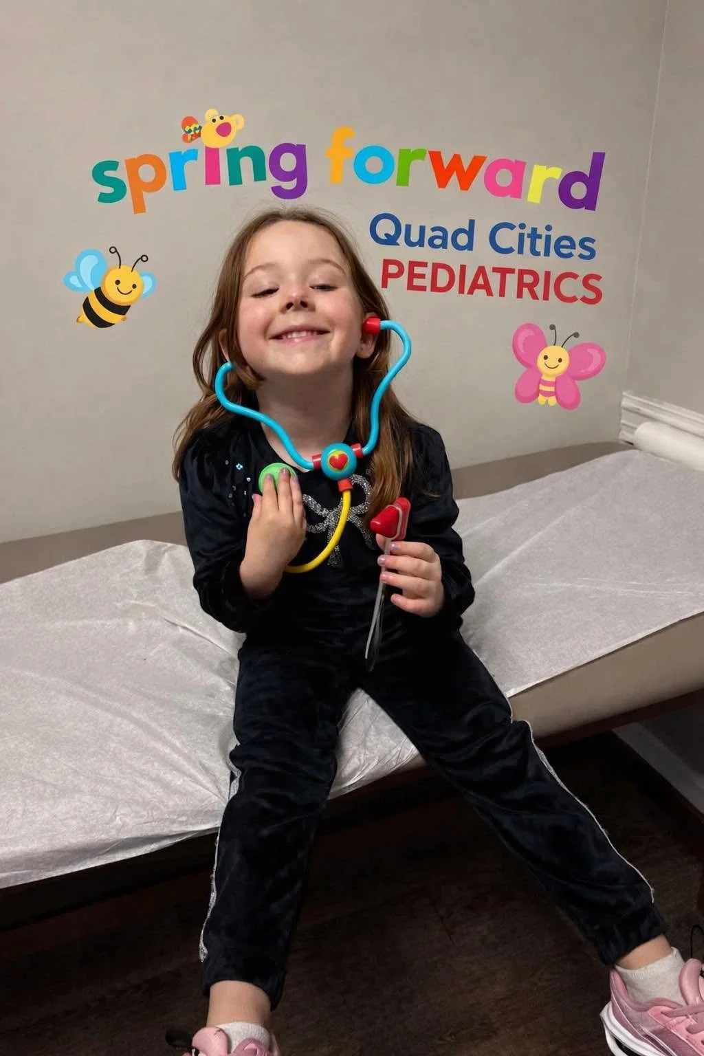 A young girl sitting on an examination bed at a pediatric clinic, holding a stethoscope, smiling, with a wall decorated with colorful foam letters and cartoon insects in the background.