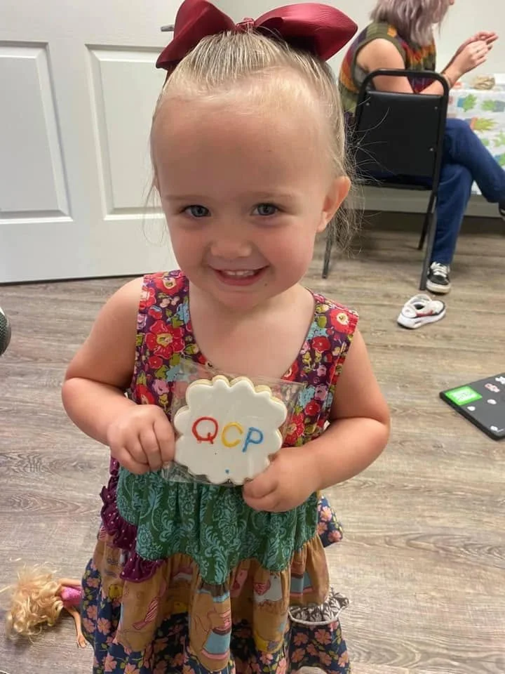 A young girl with blonde hair in a ponytail with a large red bow, smiling and holding a cookie with the letters Q, C, and P written on it in red, yellow, and blue. She is wearing a colorful dress with a floral pattern.