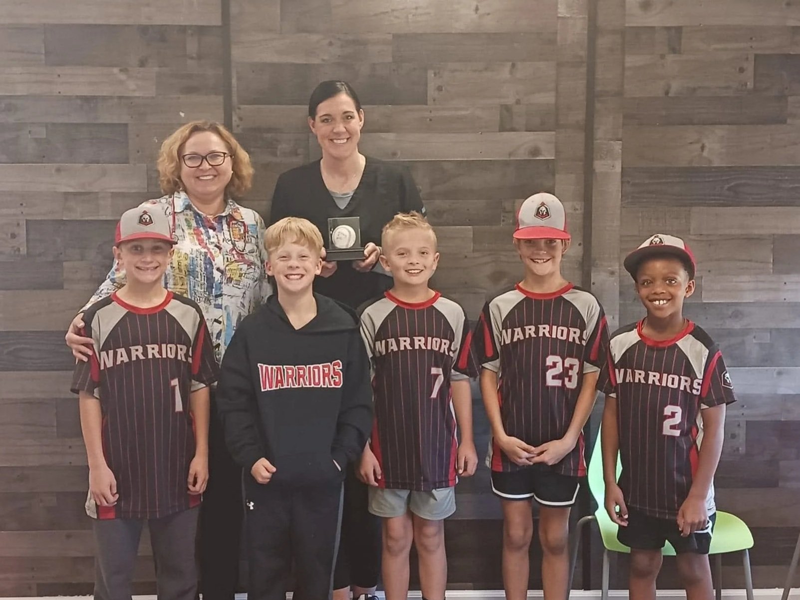 A group of five young boys in matching basketball uniforms with the word "WARRIORS" on the front, standing with two women, one of whom is holding a medal or award, against a wooden wall background.