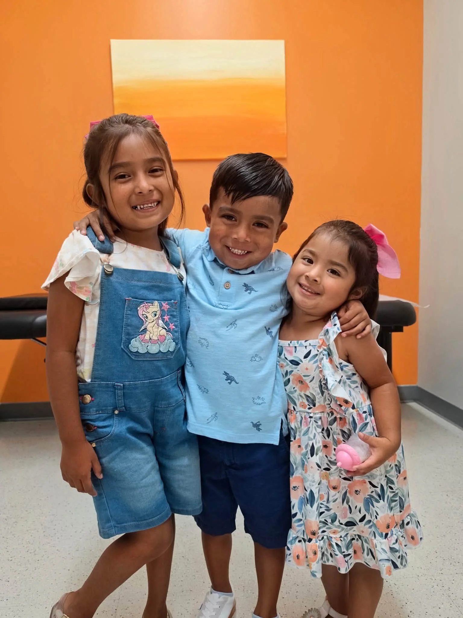 Three smiling children standing together indoors with an orange wall and a sunset painting behind them.