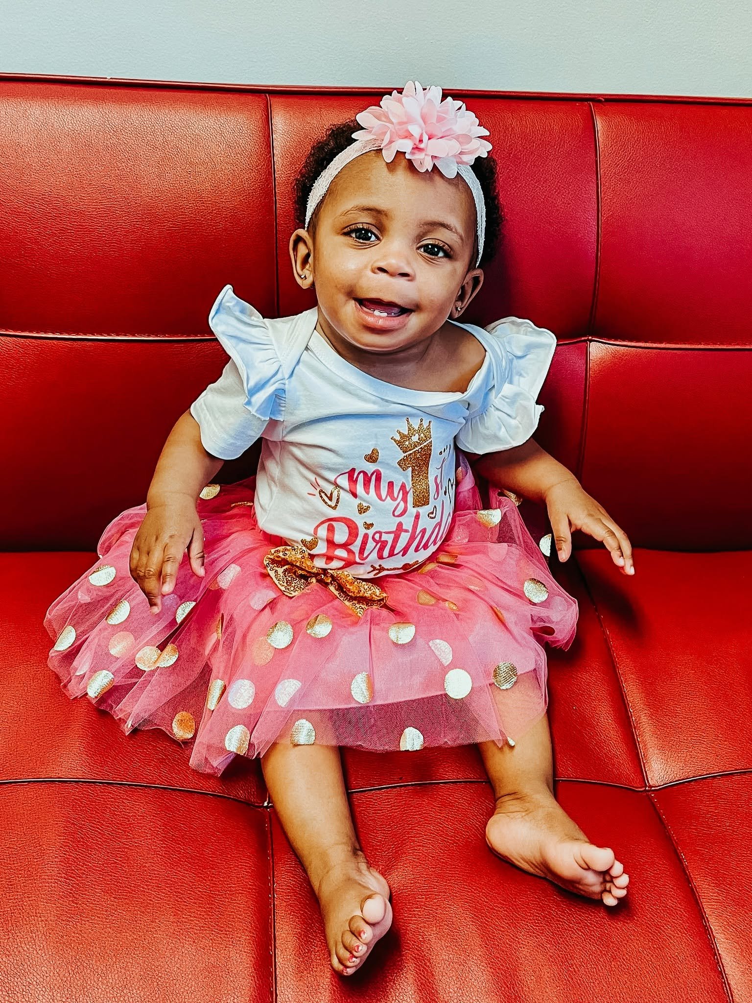A young girl sitting on a red leather couch, wearing a pink tulle skirt with gold polka dots, a white t-shirt with a birthday message, and a white headband with a pink flower. She has earrings, and her legs are extended on the couch with her bare feet visible.