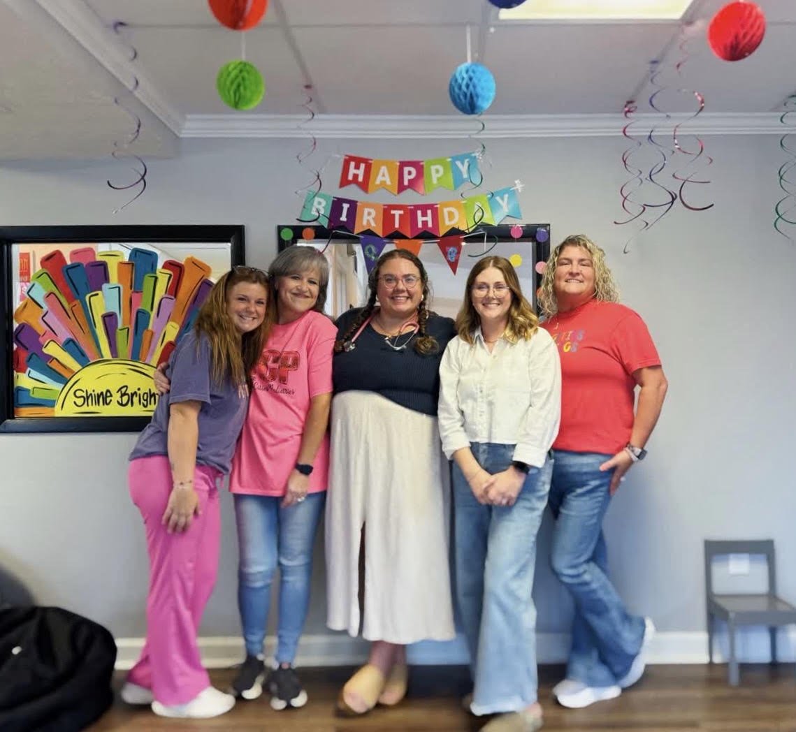 Five women standing together at a birthday party, smiling. Behind them is a colorful 'Happy Birthday' banner, hanging paper decorations, and a framed colorful artwork on the wall.