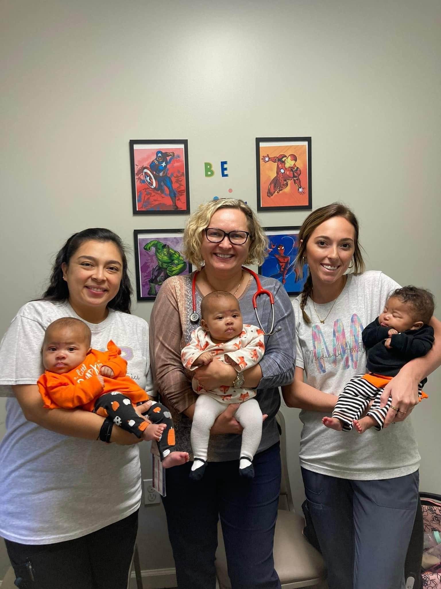 Four women standing together holding three babies, with superhero posters on the wall behind them and the words "BE" spelled out in colorful letters.