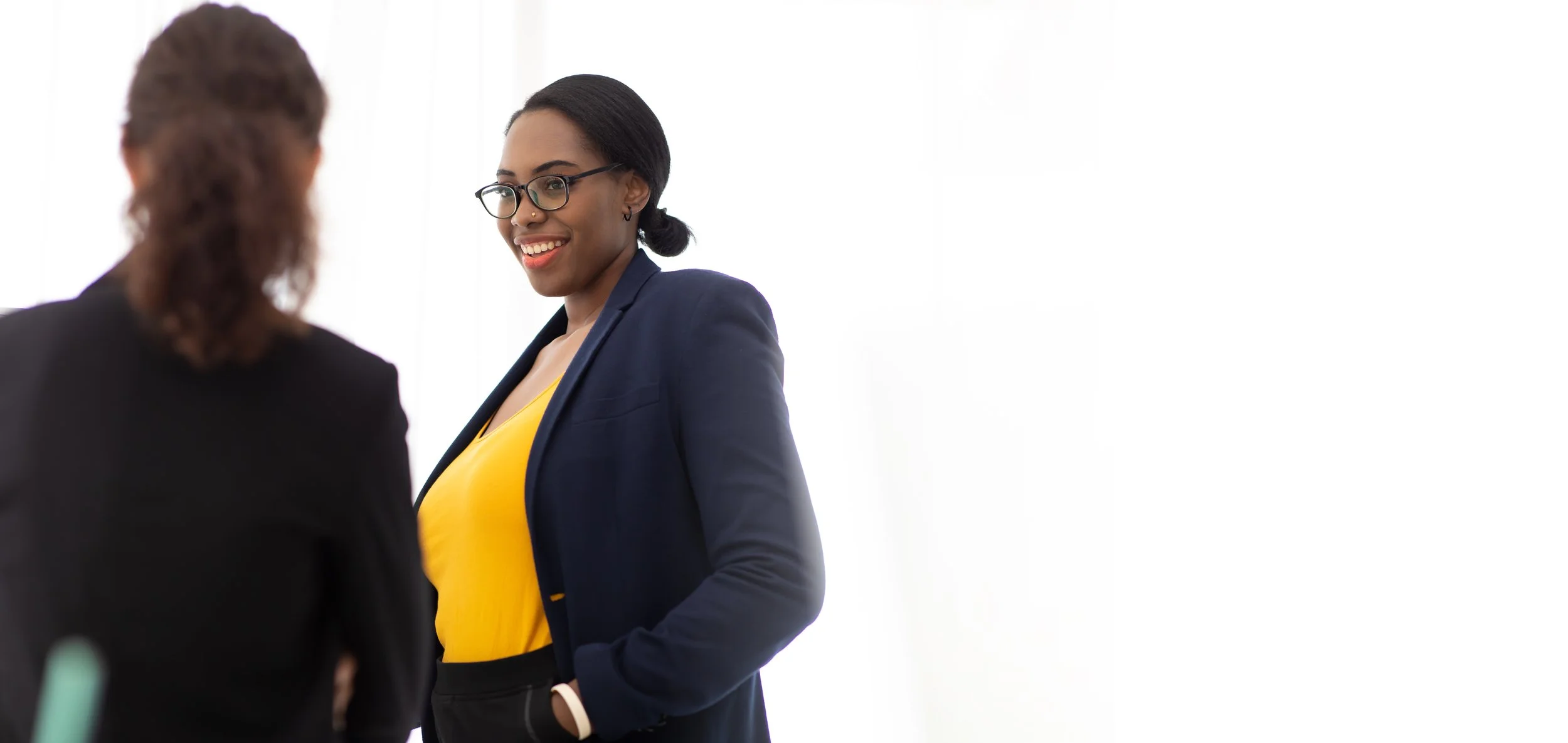 Two businesswomen talking, one smiling, wearing glasses and a blue blazer over a yellow top.