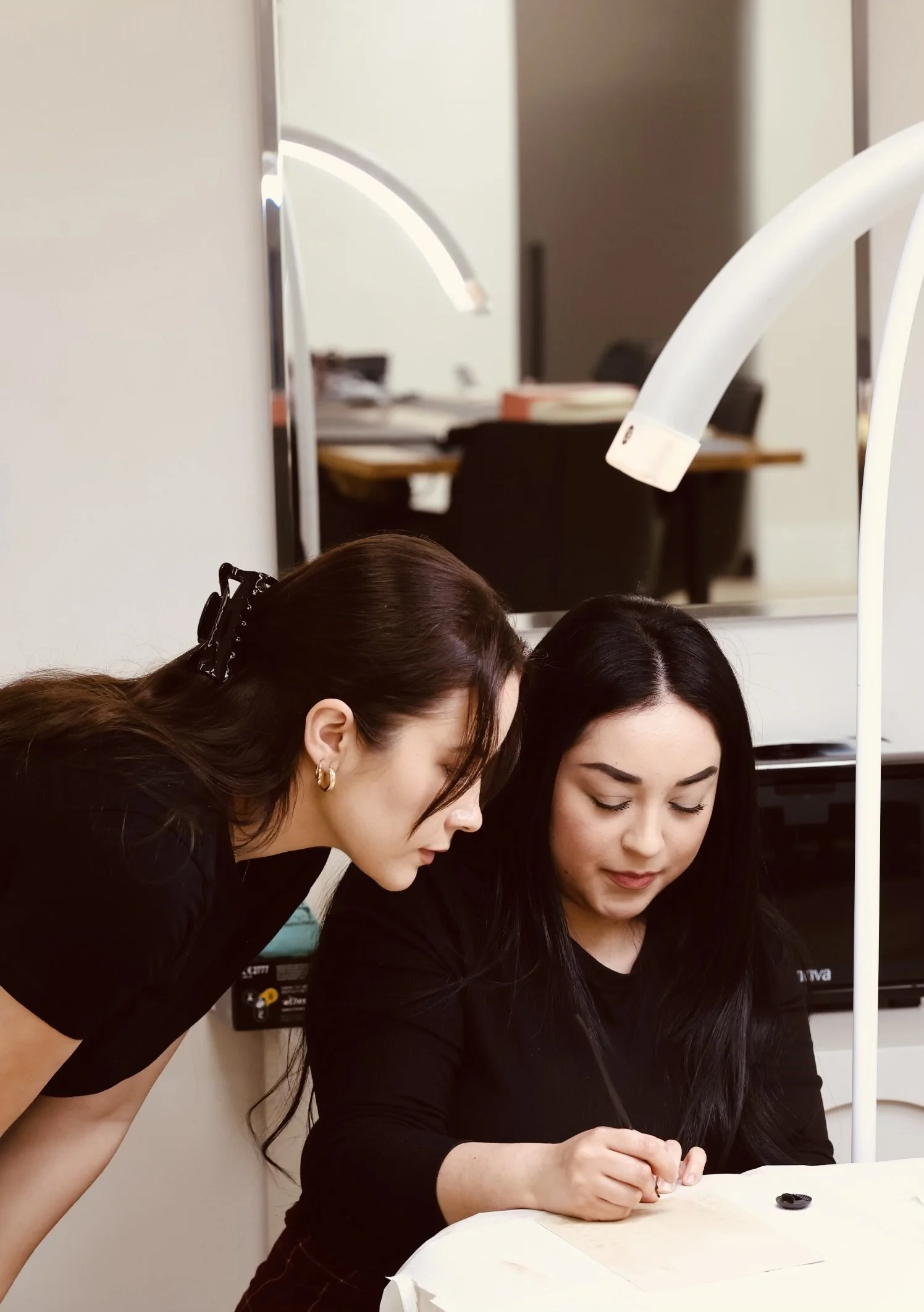 Two women working closely together at a desk, with the woman on the right writing and the woman on the left leaning in to observe.