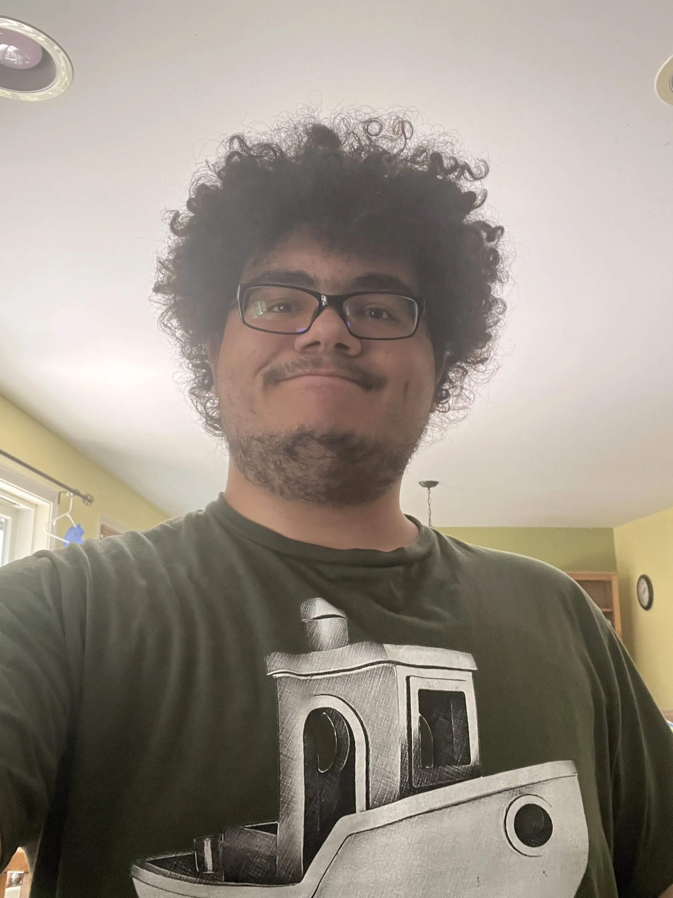 A man with curly hair and glasses taking a selfie indoors, wearing a t-shirt with a "Benchy" 3d printed boat graphic.