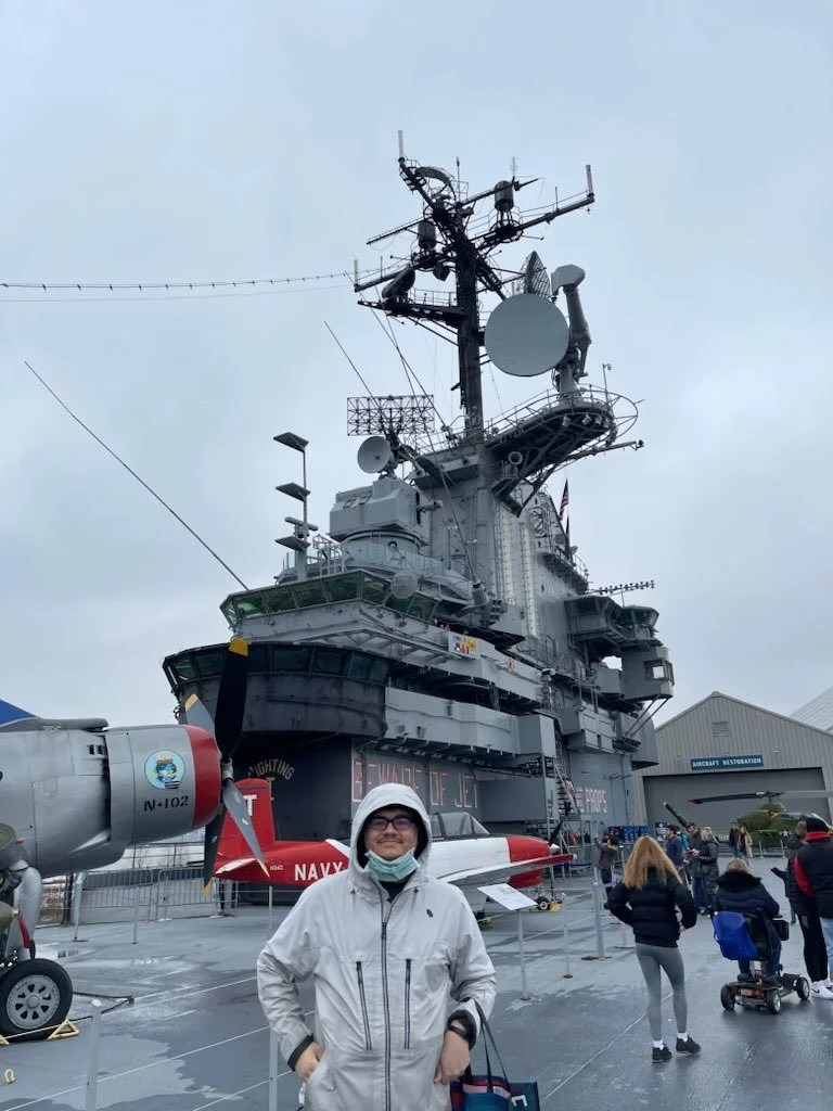 A man in a white jacket and glasses standing in front of a large military aircraft carrier with antennas and radar equipment, surrounded by people at an outdoor exhibit.
