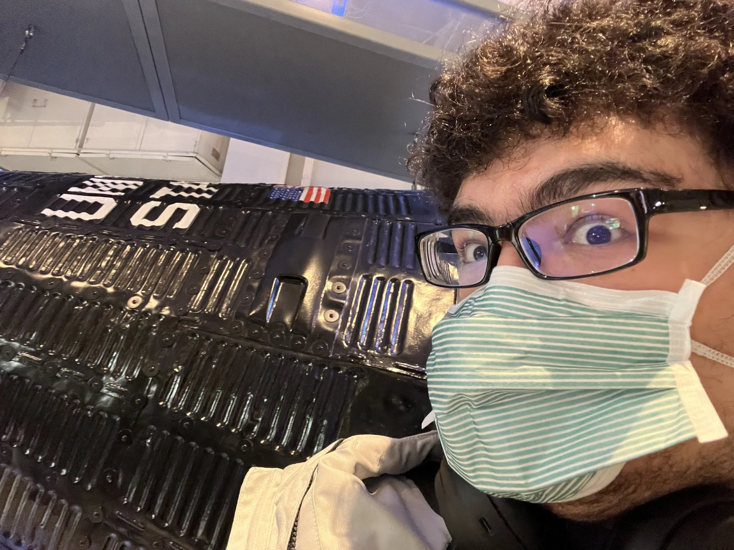A man with glasses wearing a face mask taking a selfie in front of a Mercury space capsule and an American flag.