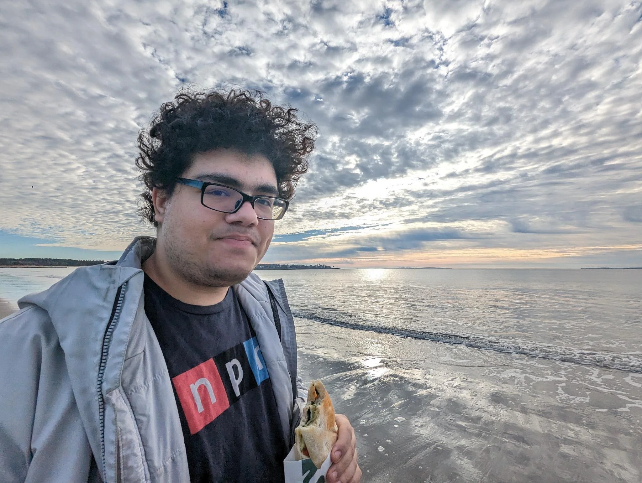 A man with curly hair, glasses, wearing a black t-shirt with a colorful graphic, and a light gray jacket, stands on a beach holding a sandwich, with the ocean and overcast sky with scattered clouds in the background.