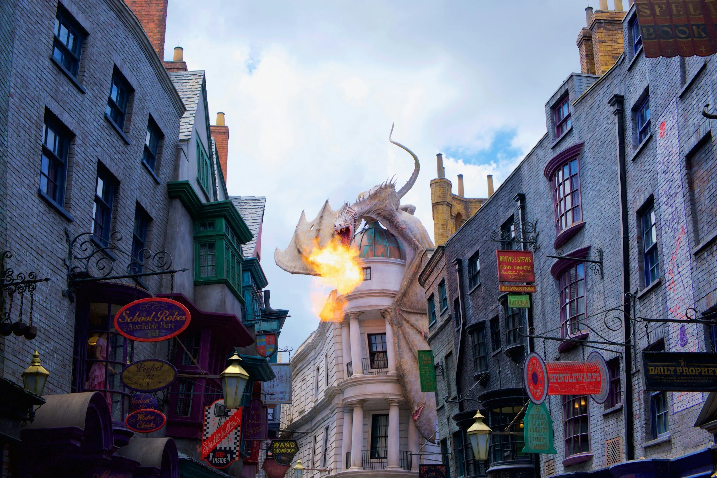 A large dragon sculpture spewing fire, perched on a building between narrow city streets with colorful storefronts and signs. In Diagon Alley at Universal Studios Theme Park in Universal Orlando Resort in Orlando, FL.