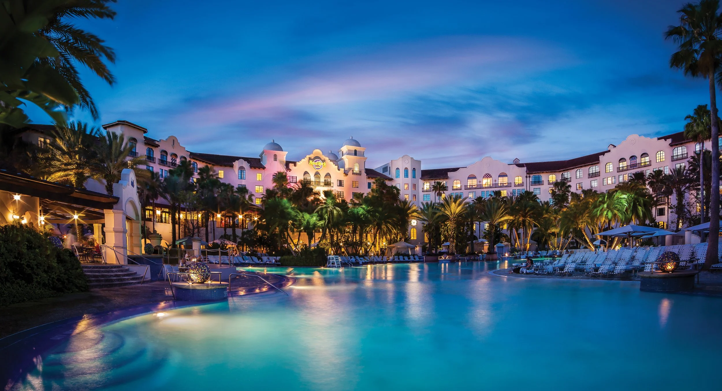Nighttime view of a luxurious resort with a large swimming pool, surrounded by palm trees, lounge chairs, and a multi-story hotel building with illuminated windows and a visible Hard Rock Cafe sign.