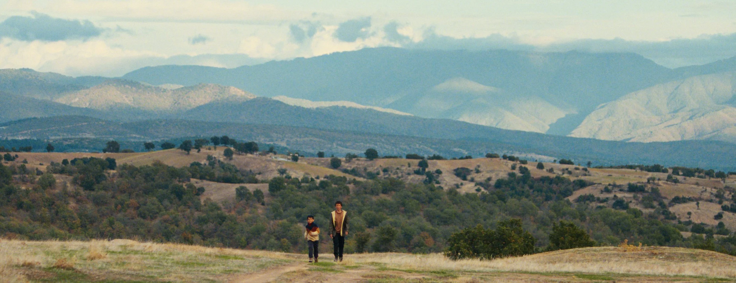 Two boys walking in a grassy field with rolling hills and mountains in the background during daytime.