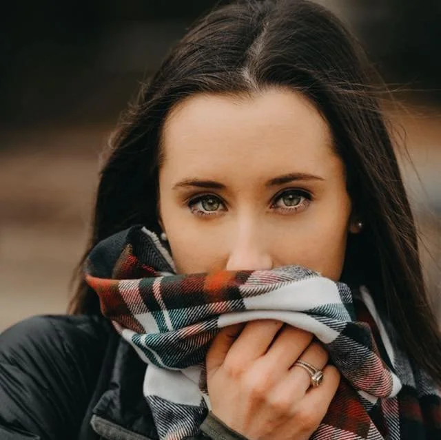 A woman with long dark hair and blue eyes is holding a multicolored plaid scarf over her mouth and nose, wearing a black jacket, outdoors.