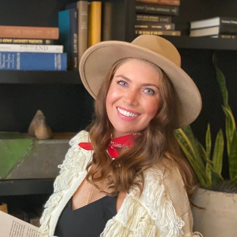 A woman with long wavy brown hair, wearing a large beige hat, black top, red bandana around her neck, and a cream-colored textured jacket, smiling in front of a bookshelf.