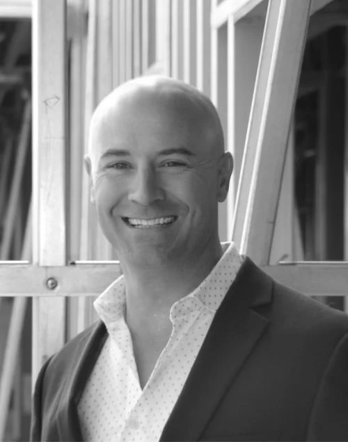 A smiling man with a shaved head wearing a suit jacket and white polka dot dress shirt, standing in front of a metal framework.