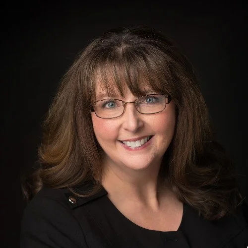 Headshot of a woman with shoulder-length brown hair, glasses, and a black top, smiling against a dark background.