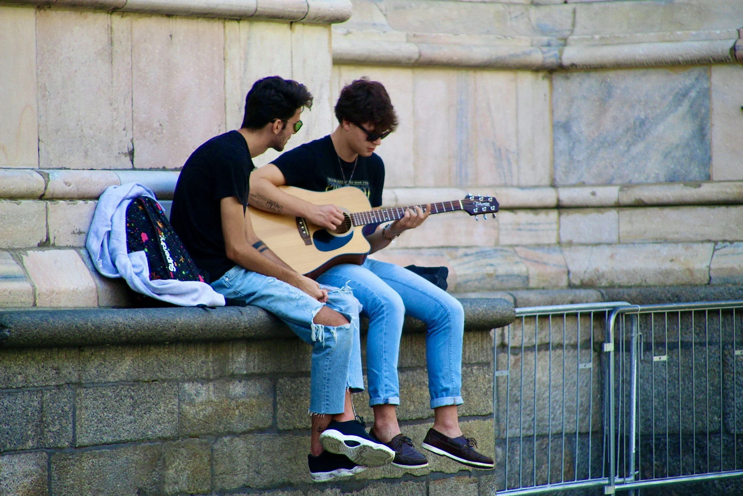 Two young men sitting on a stone bench outdoors, one playing an acoustic guitar. One has sunglasses and the other is wearing ripped jeans. There is a backpack and a hoodie beside them.