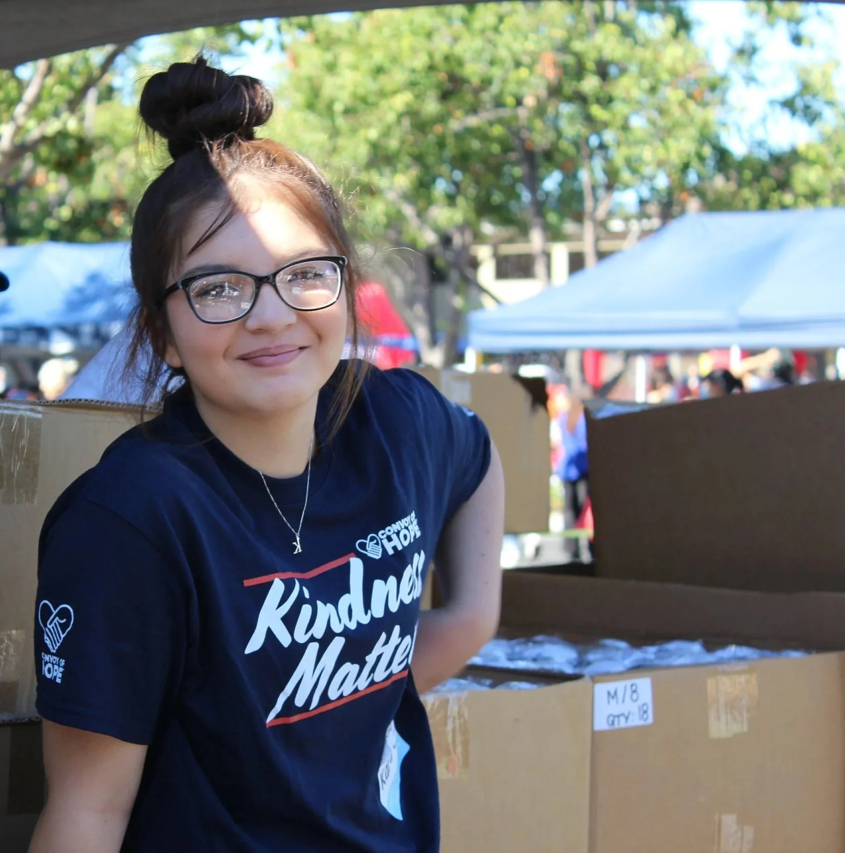 A young woman with glasses and a black t-shirt that reads 'Kindness Matters' stands outdoors at a community event, surrounded by tents and boxes.