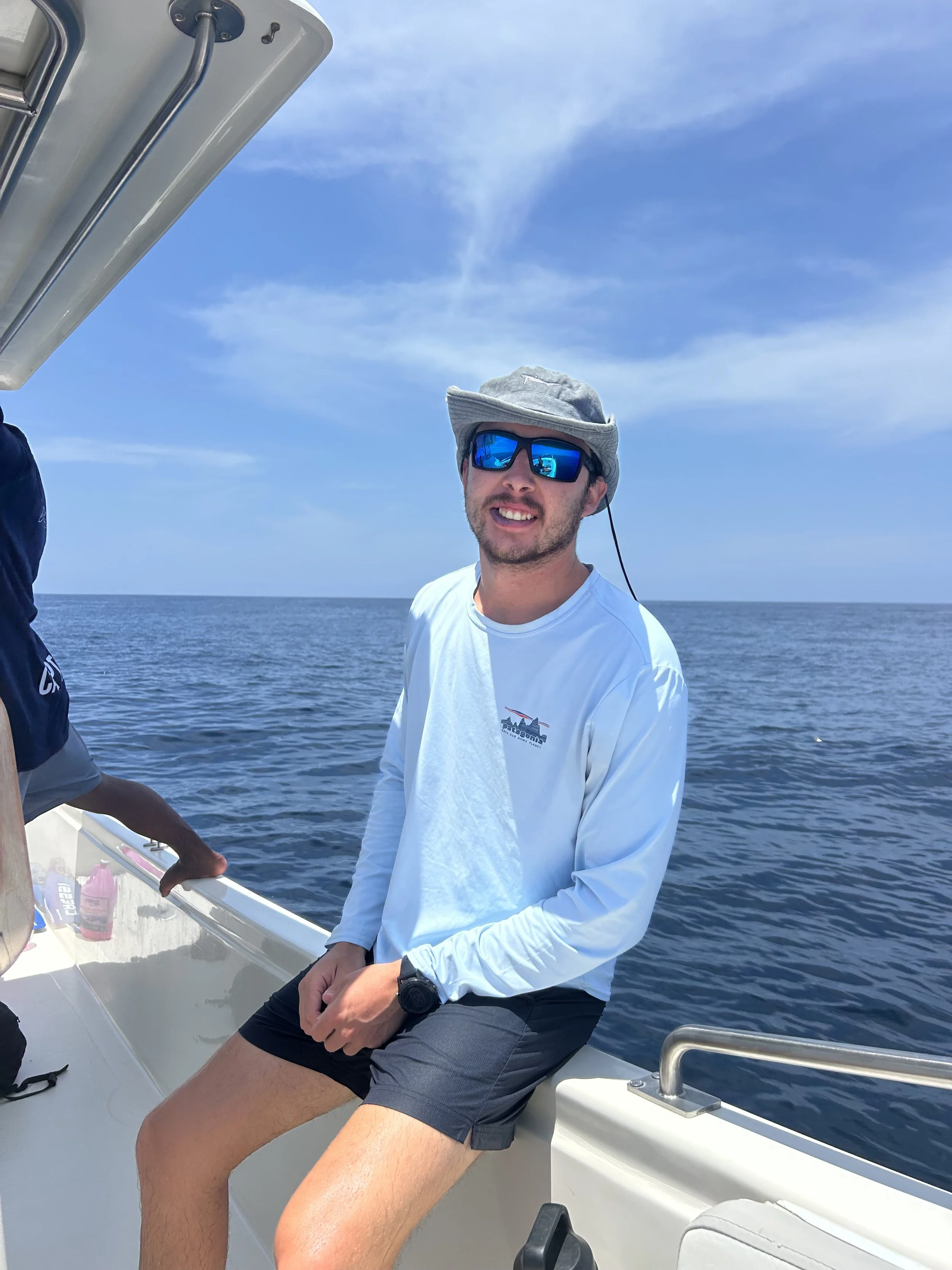 Person wearing sunglasses and a white T-shirt, smiling outdoors near a body of water, with palm trees in the background.