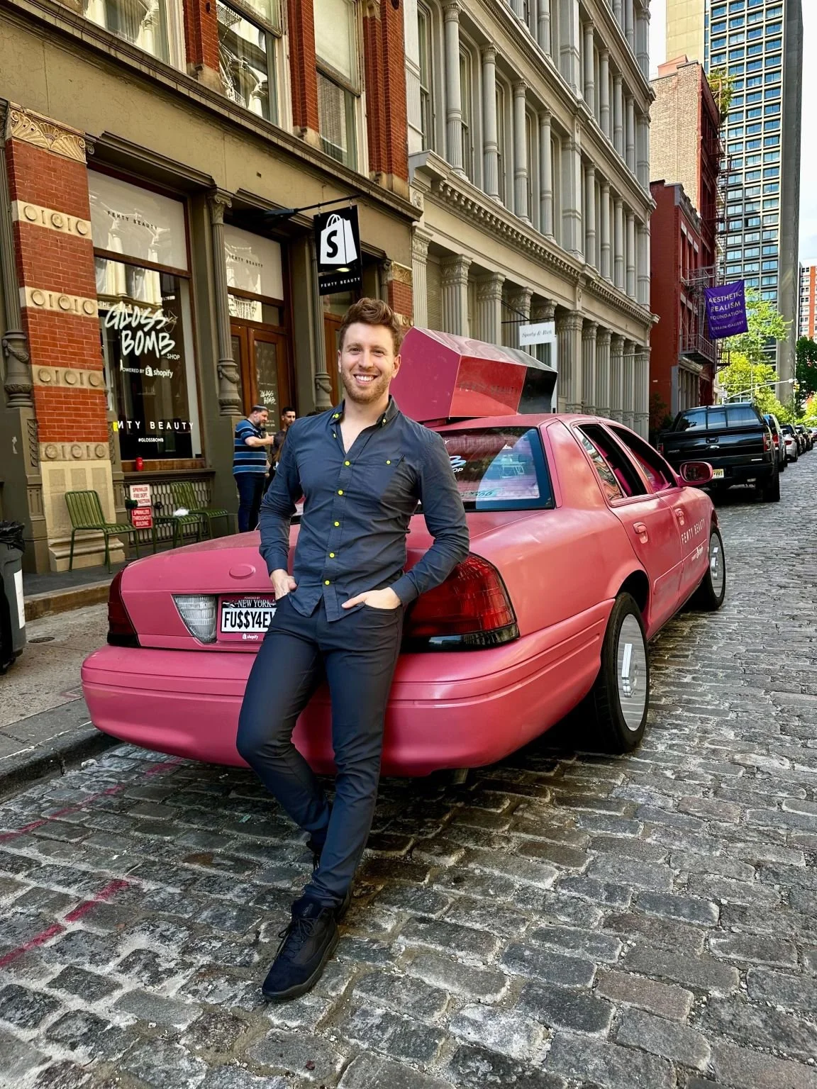 A smiling man in a dark shirt and black pants stands with hands in pockets in front of a Fenty Beauty pink car with a custom rear hatch on a cobblestone street in SOHO, New York, with historic buildings and storefronts in the background.