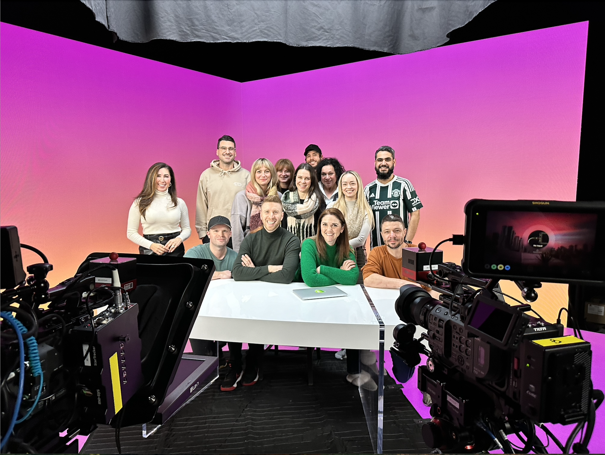 Group of diverse people smiling and posing for a photo in front of a colorful pink gradient background, with professional video cameras and equipment in the foreground.