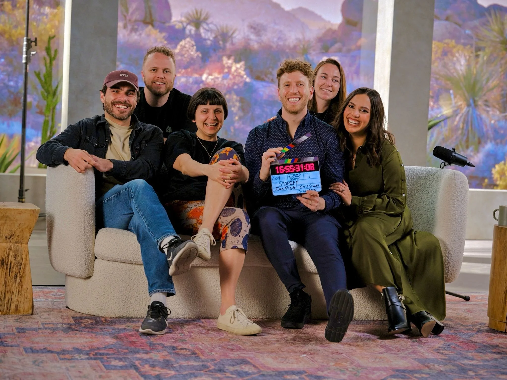A group of six smiling people sitting on a sofa on a studio set with a colorful abstract background. One person in the front is holding a clapperboard. There is a microphone on a stand next to them.