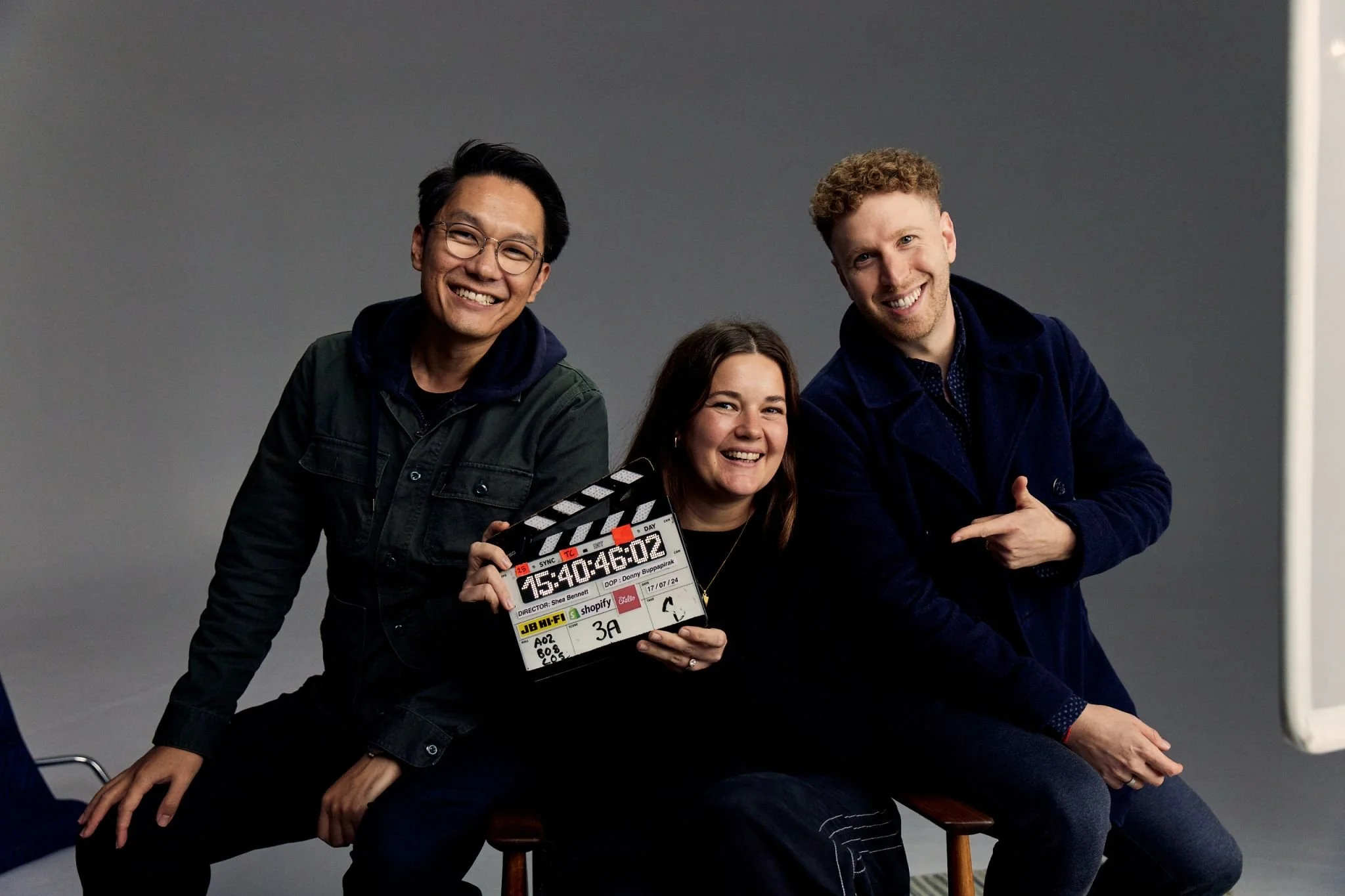Three smiling people, two men and one woman, posing with a film clapboard in front of a plain gray background.