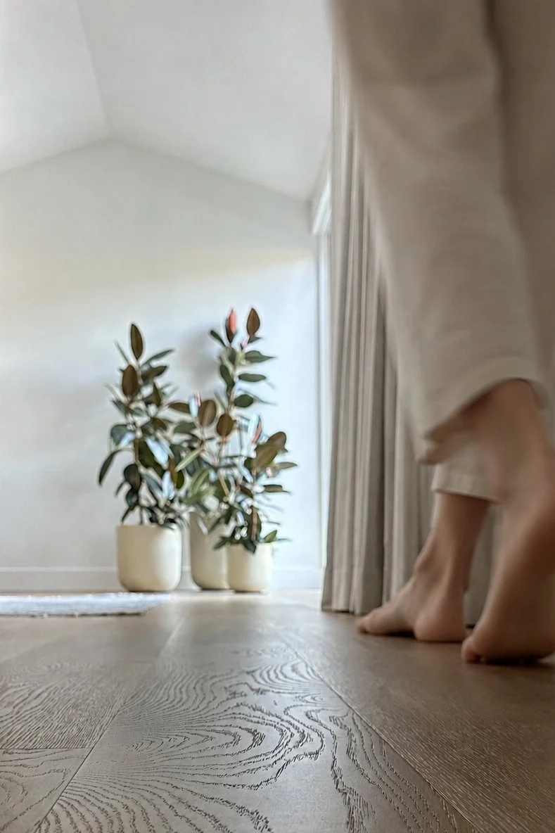 Interior of a room with a wooden floor, a large potted plant with broad leaves, and beige curtains next to a person standing barefoot with only lower legs and feet visible.