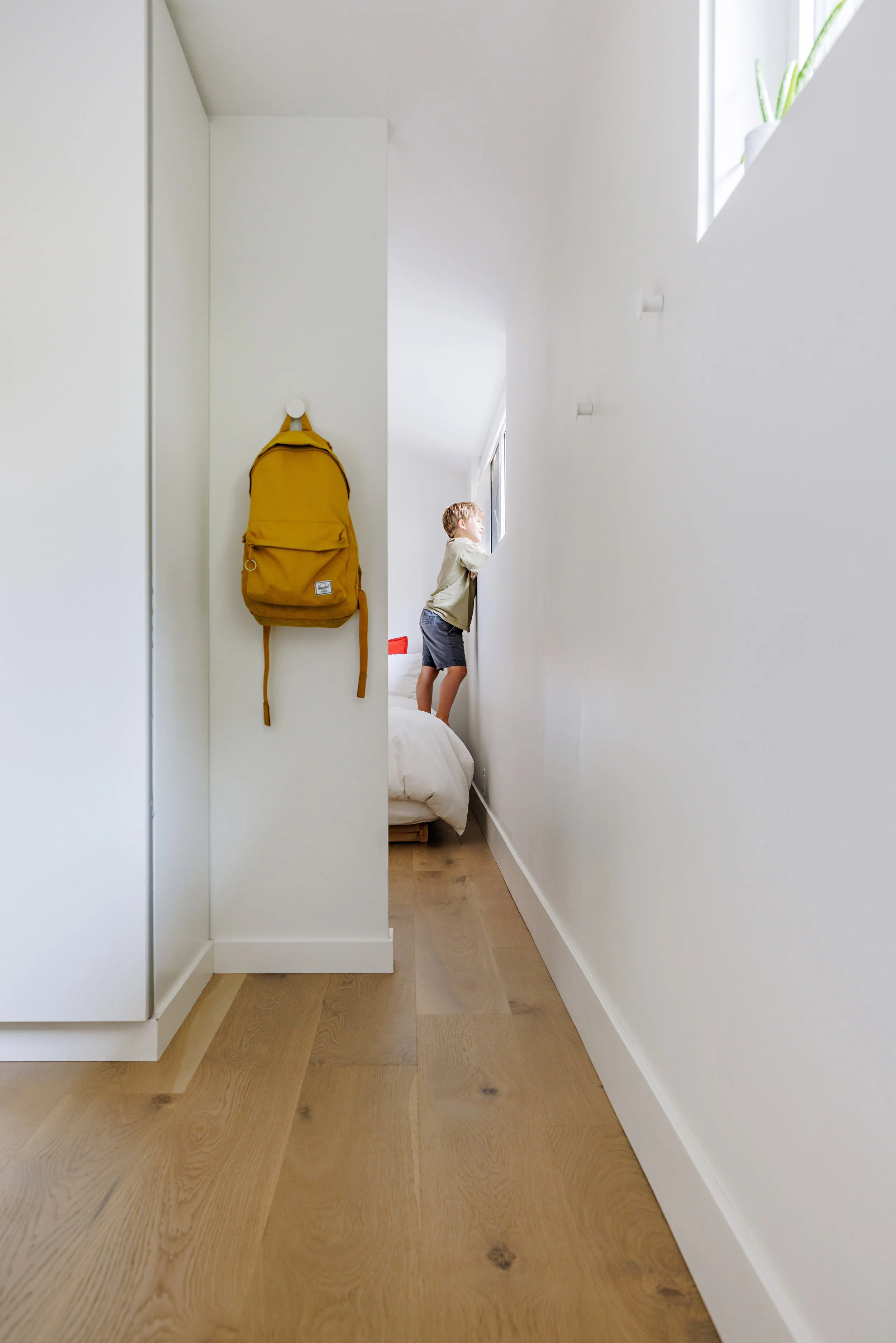 A minimalistic interior with light wooden flooring, white walls, a small bed partially visible, a yellow backpack hanging on a white wall, and a young boy looking out a window.