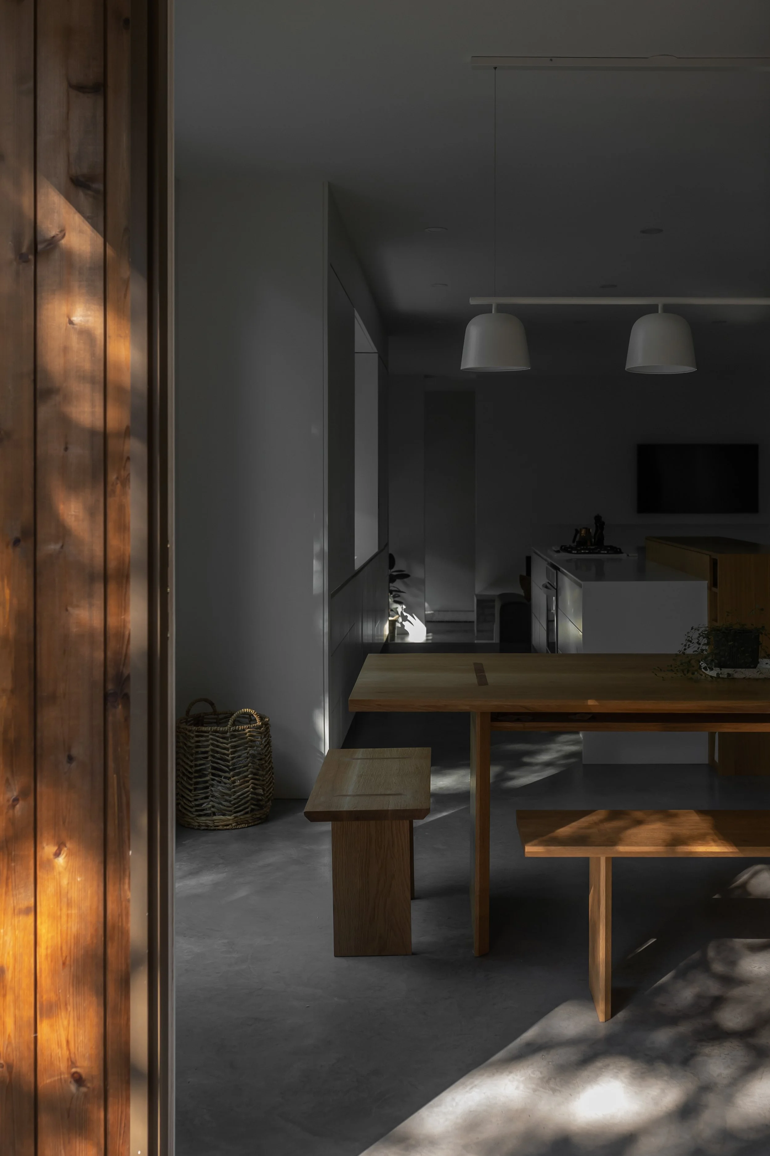 Interior of a modern home with a wooden dining table, bench, and woven basket, illuminated by natural light and featuring minimalist design elements.